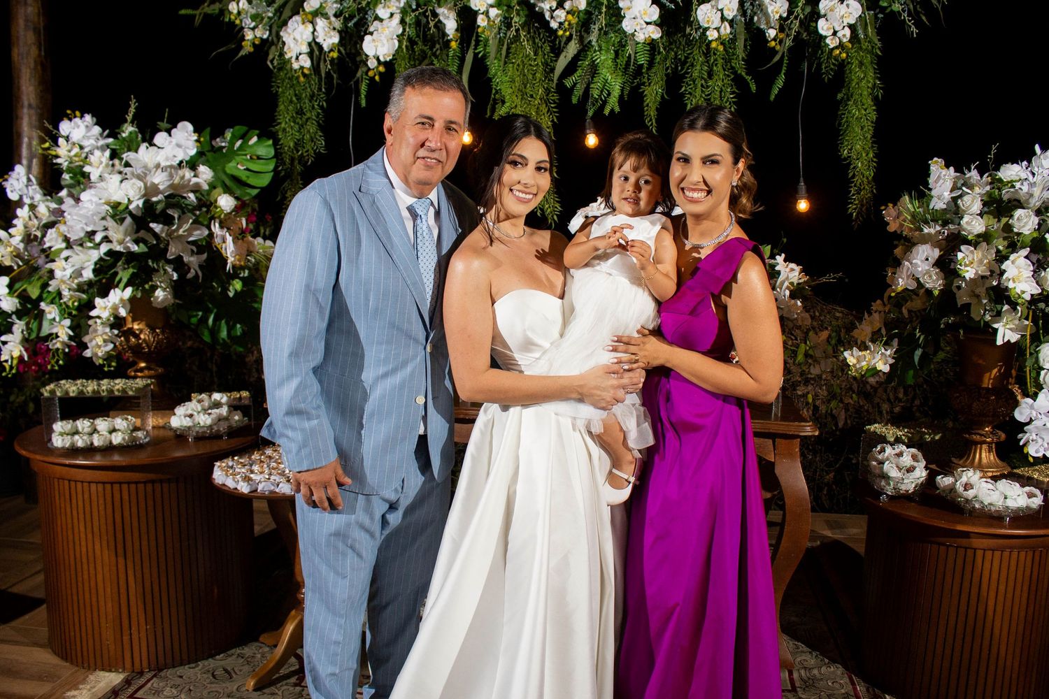 Bride in white gown poses with family including man in blue suit, woman in purple dress, and child at floral wedding reception.