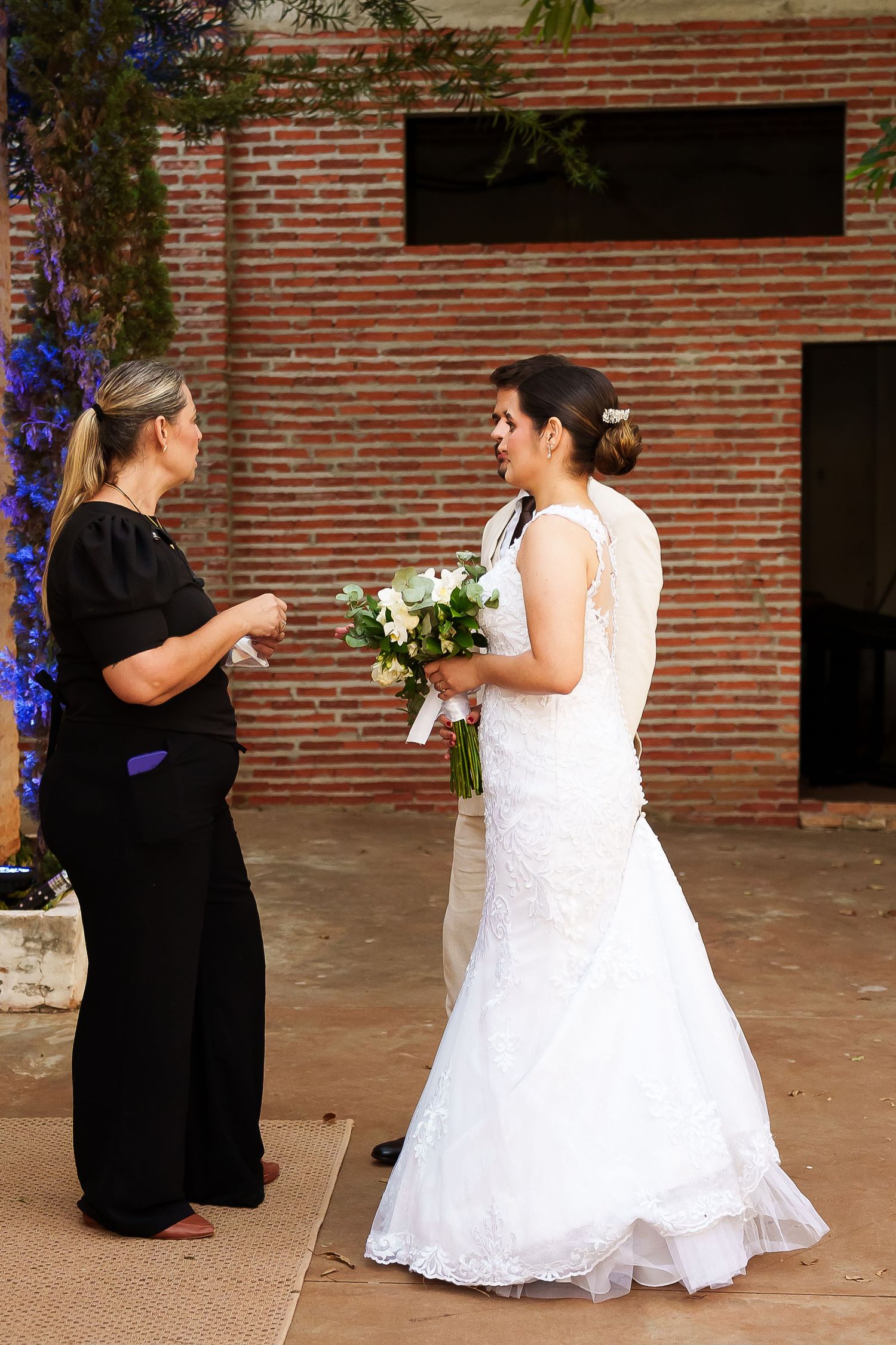 Bride in white lace gown holding bouquet conversing with woman in black outfit at outdoor wedding venue.