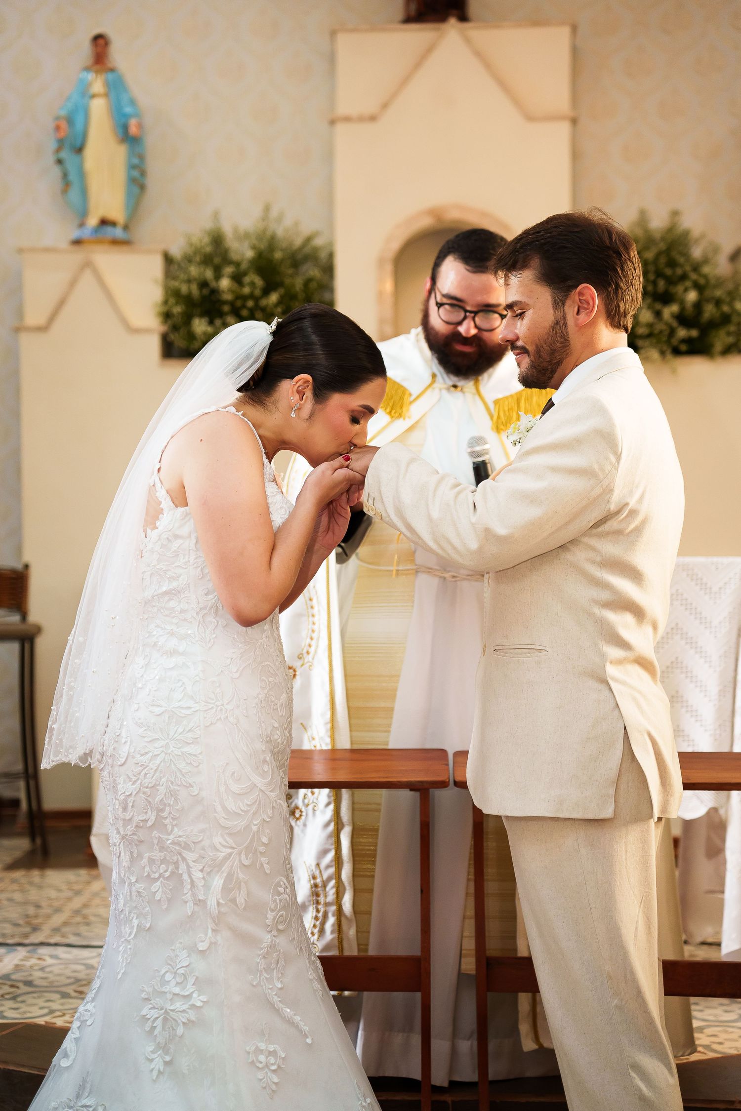 Bride kissing groom's hand during Catholic wedding ceremony at church altar with priest officiating.