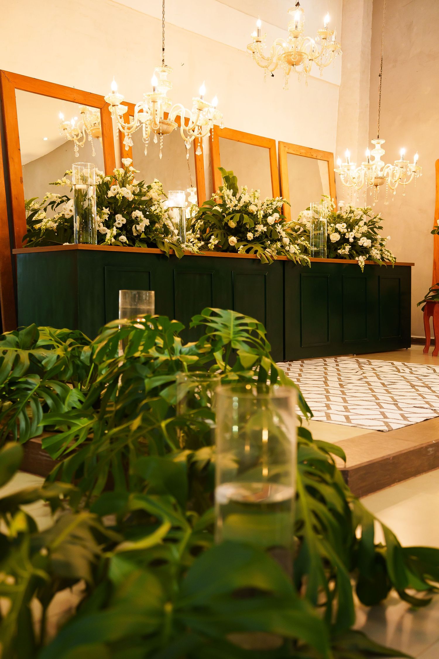 Elegant event space with green cabinetry, floral arrangements, gold-framed mirrors, and crystal chandeliers.