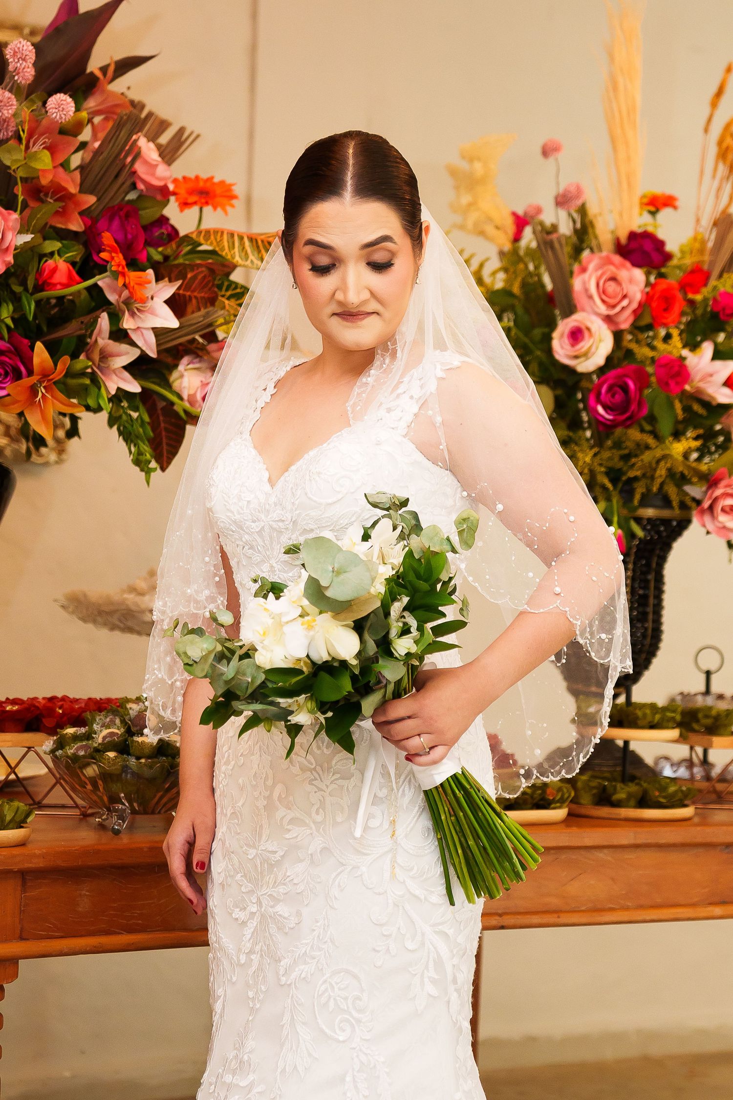 Bride in white lace gown and veil holds white bouquet, standing before colorful floral arrangements at wedding ceremony.