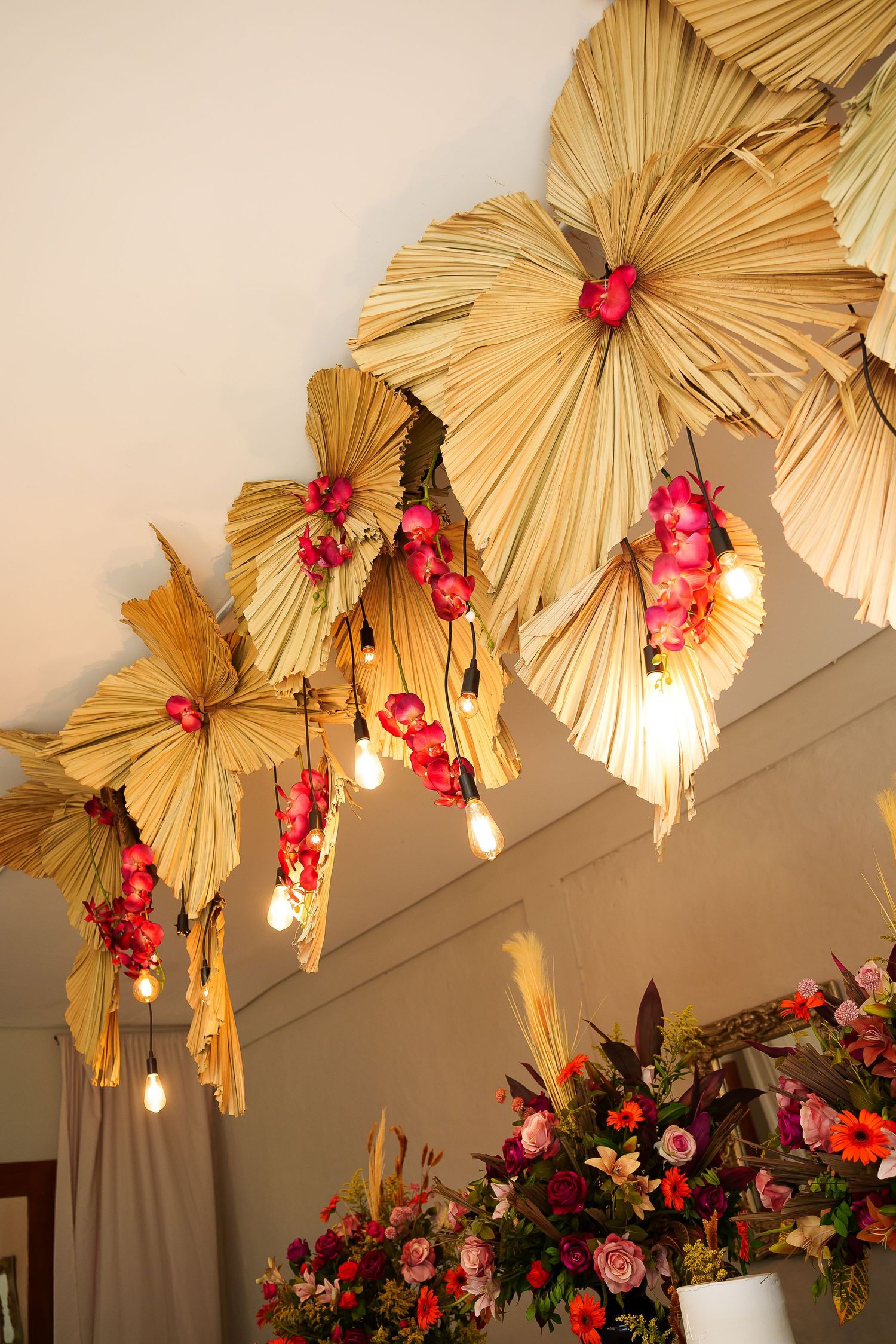 Dried palm leaf fans with pink flowers and Edison bulbs hang from ceiling above colorful floral arrangements.