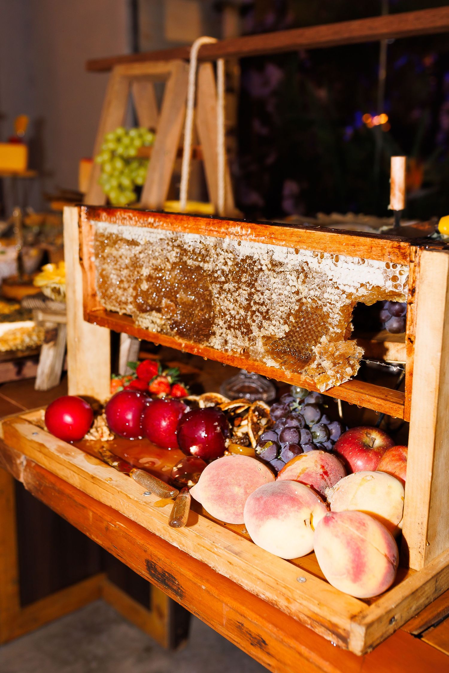 Wooden display with honeycomb frame, fresh peaches, red apples, and grapes at a rustic food market.