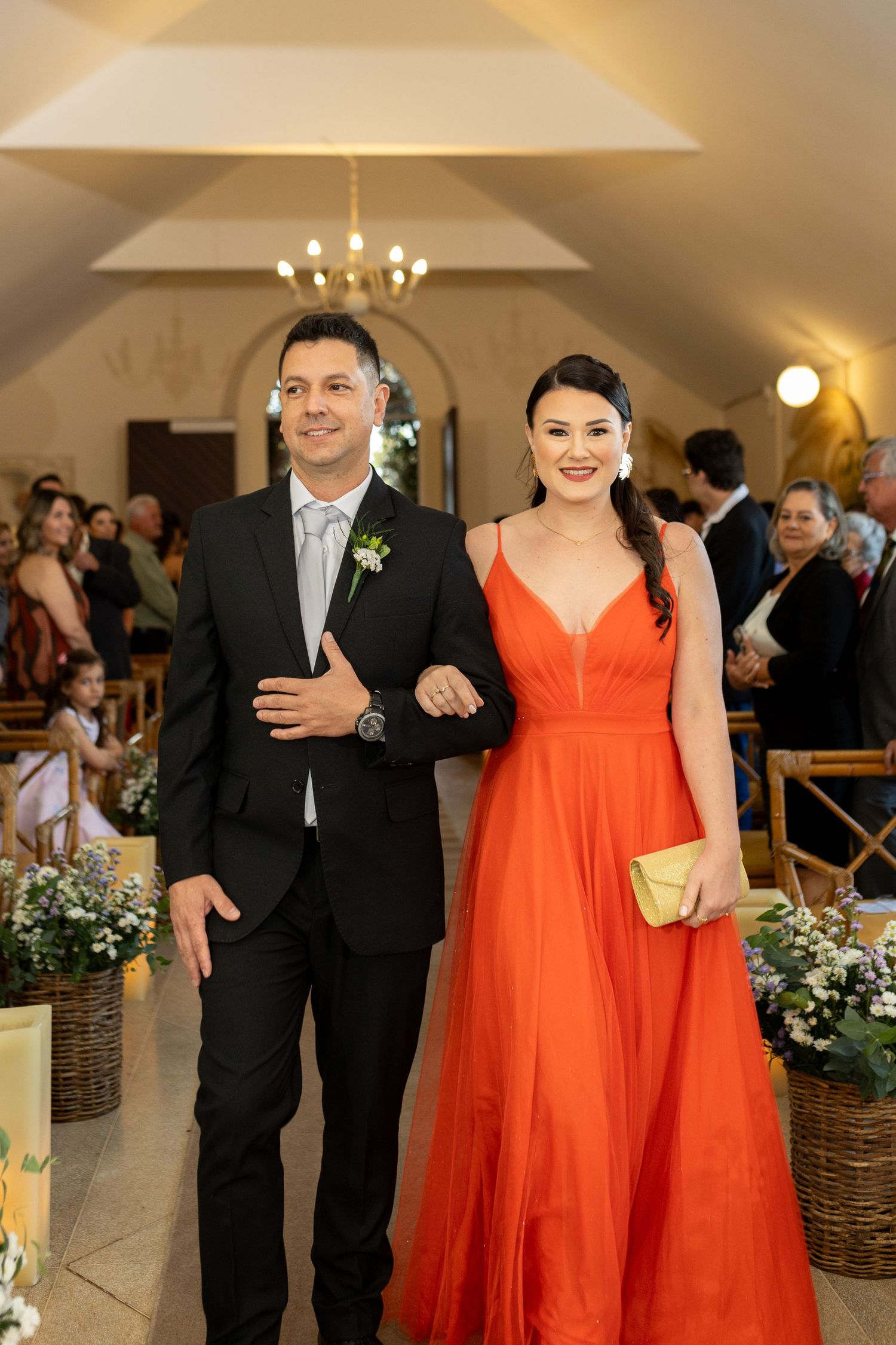 Smiling couple in formal attire walk down decorated church aisle, woman in orange gown at wedding.