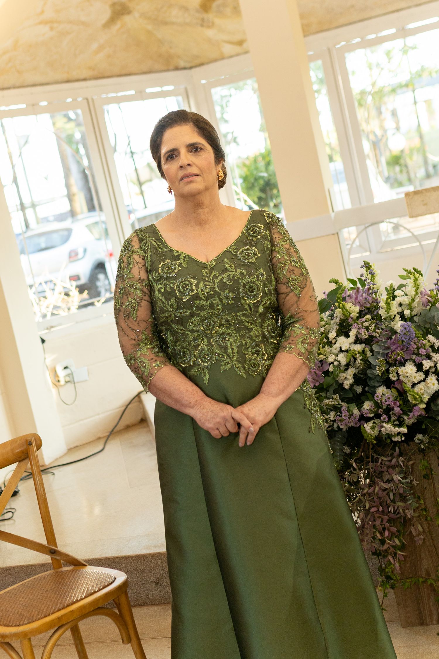 Elegant woman in olive green embroidered gown standing in decorated venue with floral arrangements.