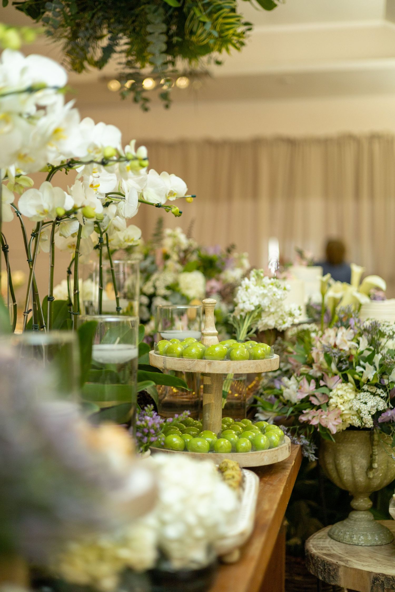 Elegant wedding dessert table with green macarons, white orchids, and lush floral arrangements in soft, romantic lighting.
