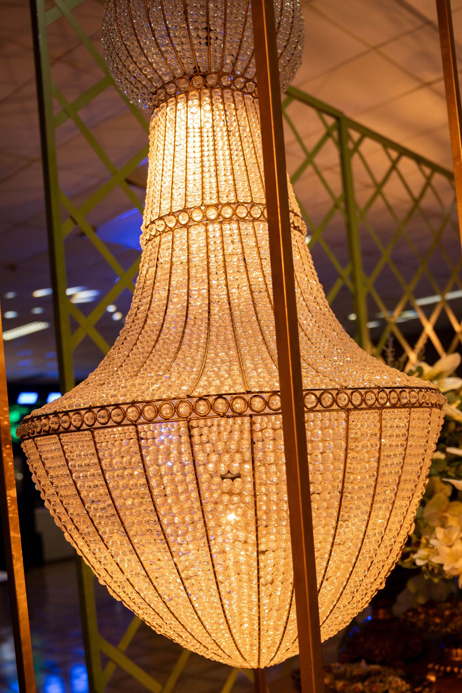 Elegant crystal beaded chandelier glowing warmly, hanging in a decorated venue with green lattice backdrop.