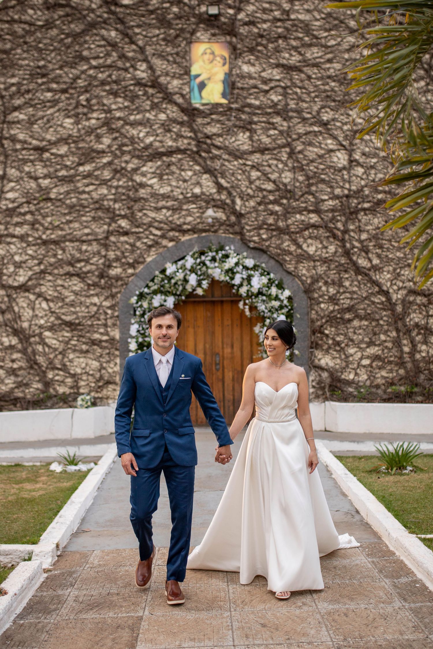 Bride and groom walk hand in hand from a vine-covered chapel with floral arch, she in white gown, he in blue suit.