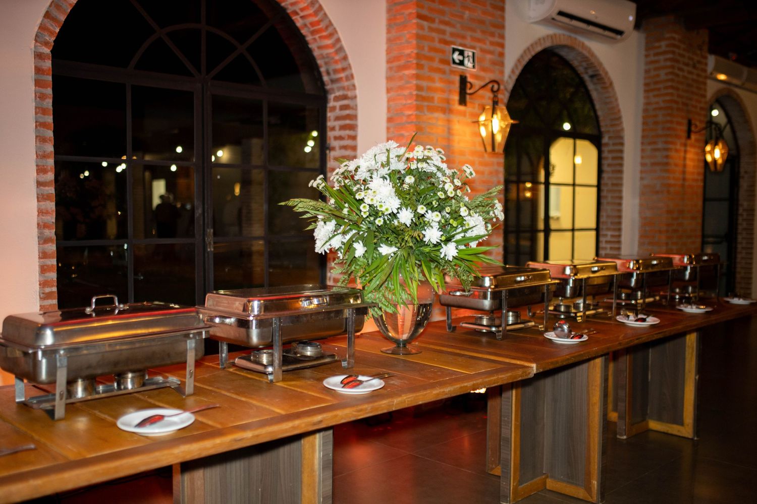 Elegant buffet setup with chafing dishes and floral centerpiece on wooden table in rustic brick restaurant.