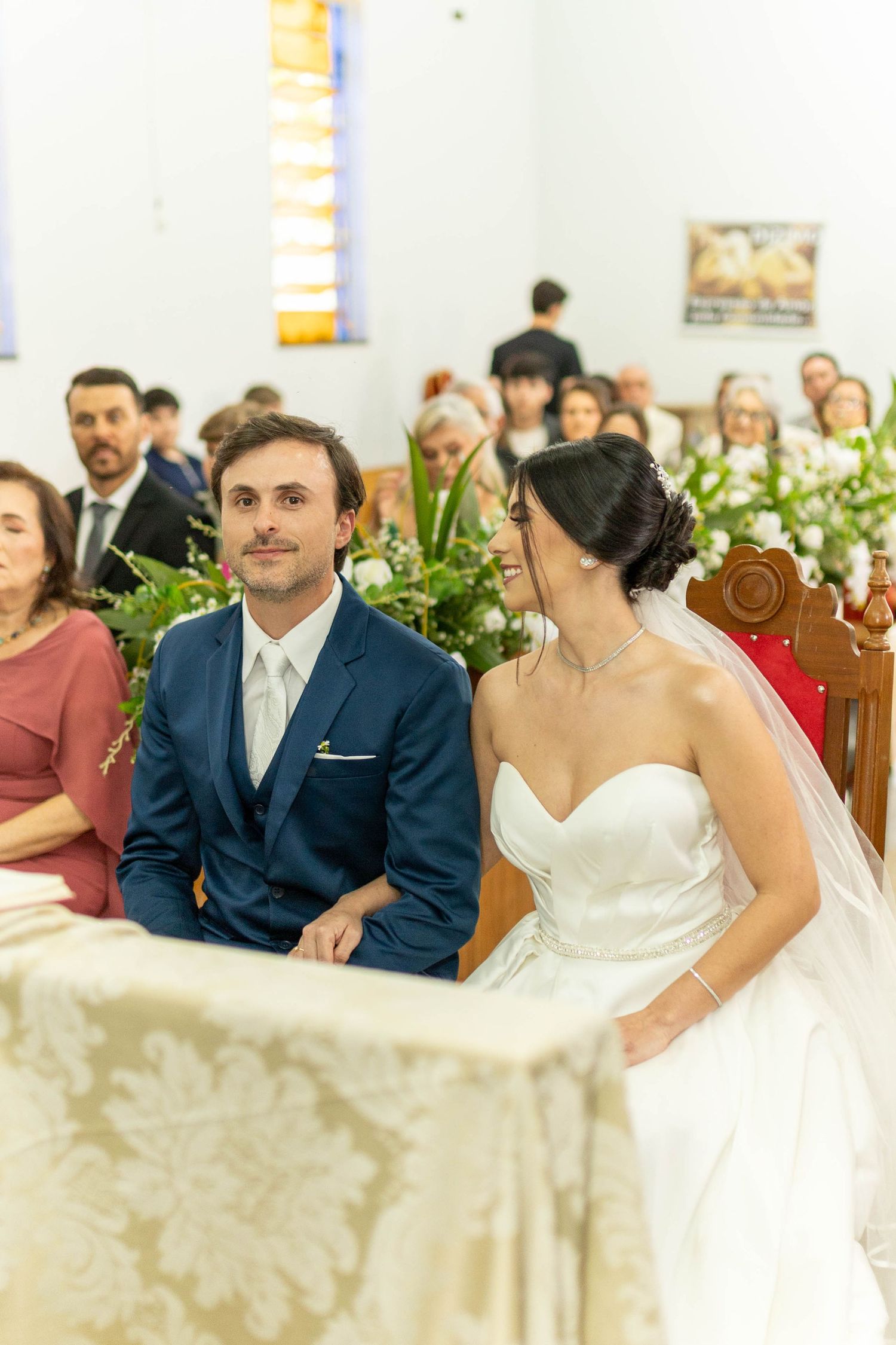 Bride and groom seated at altar during church wedding ceremony, surrounded by floral arrangements and guests.