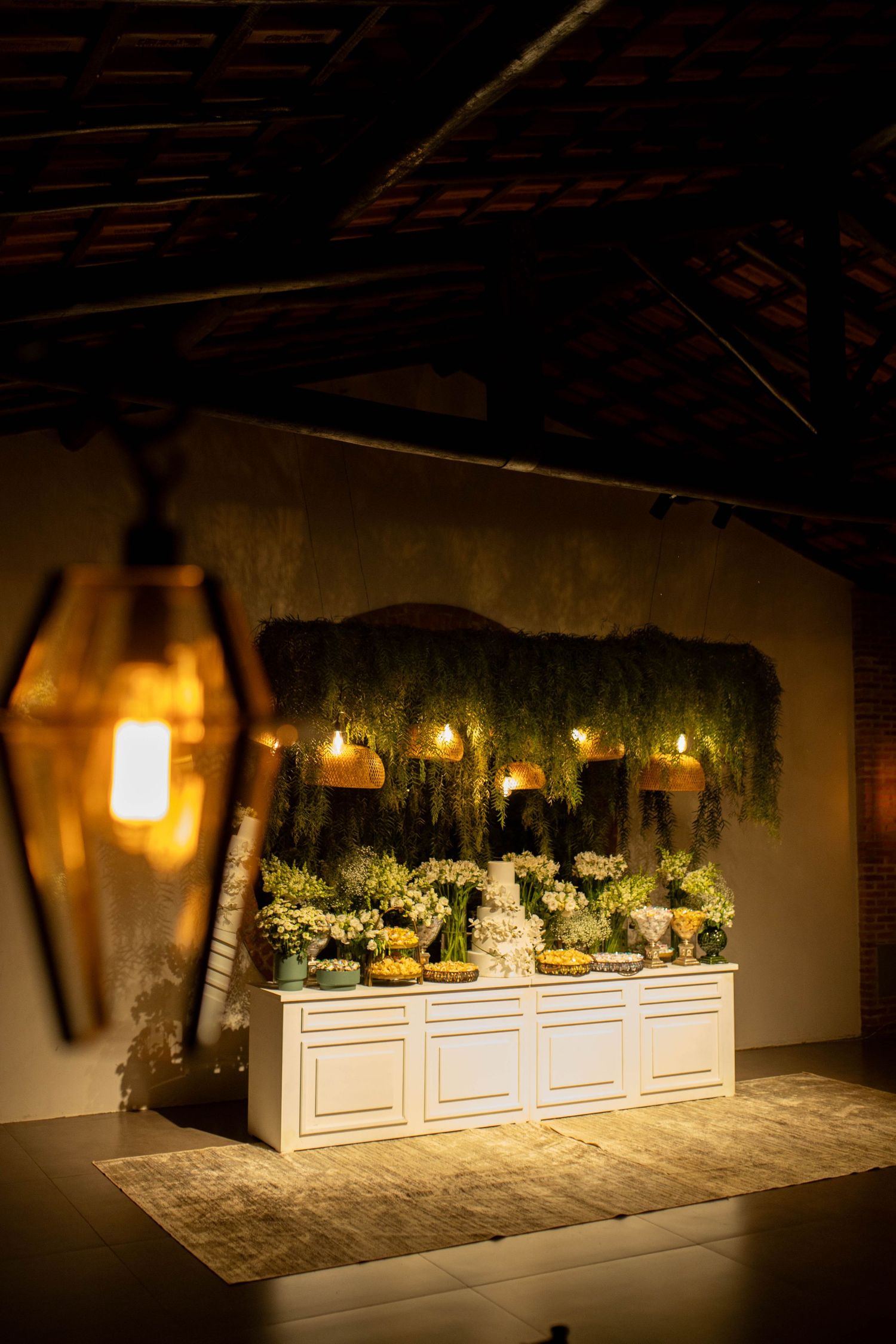 Elegant wedding dessert table with white cabinetry, lush greenery backdrop, candles, and floral arrangements in warm ambient lighting.