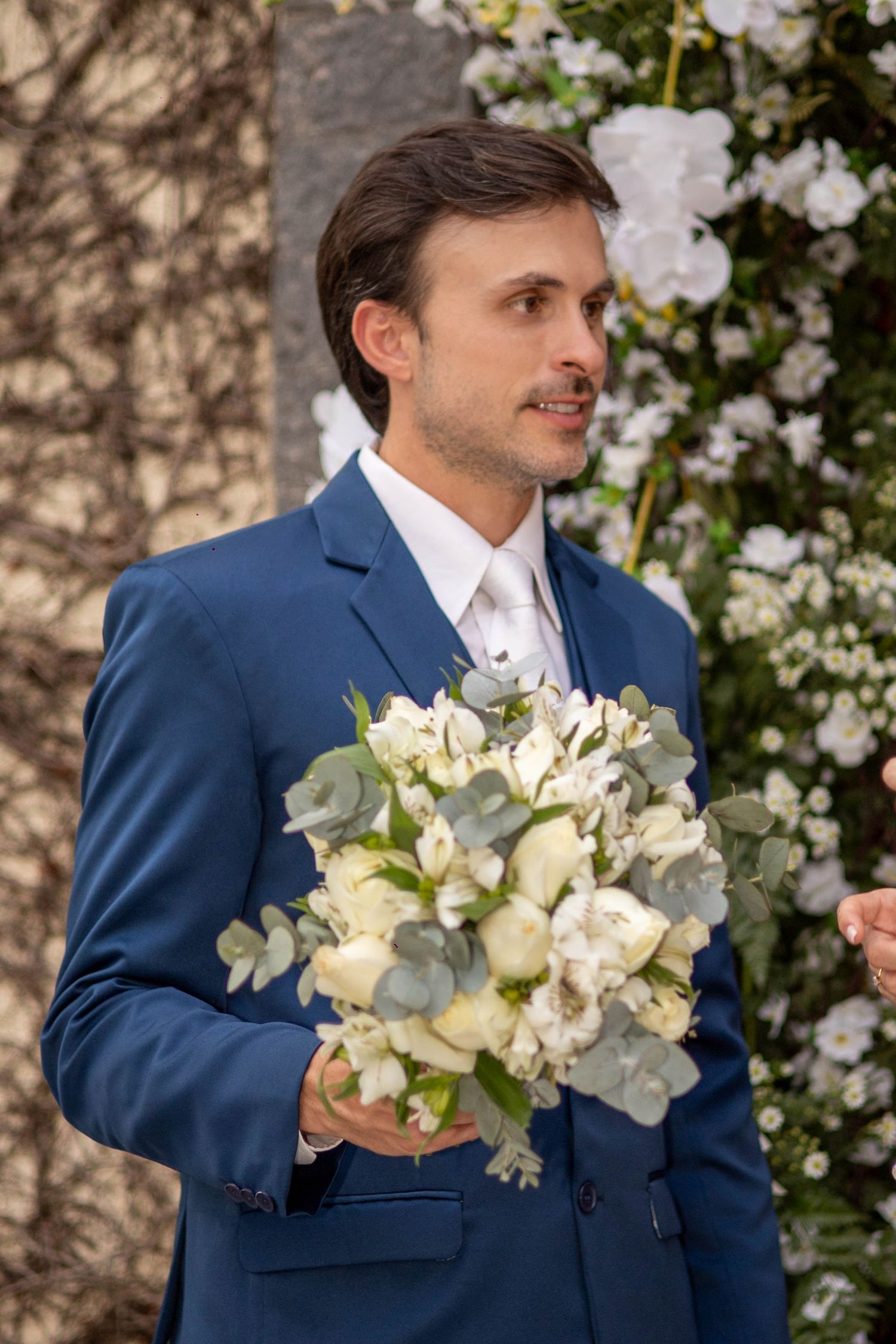 Groom in blue suit holding white floral bouquet with eucalyptus at wedding ceremony outdoors.