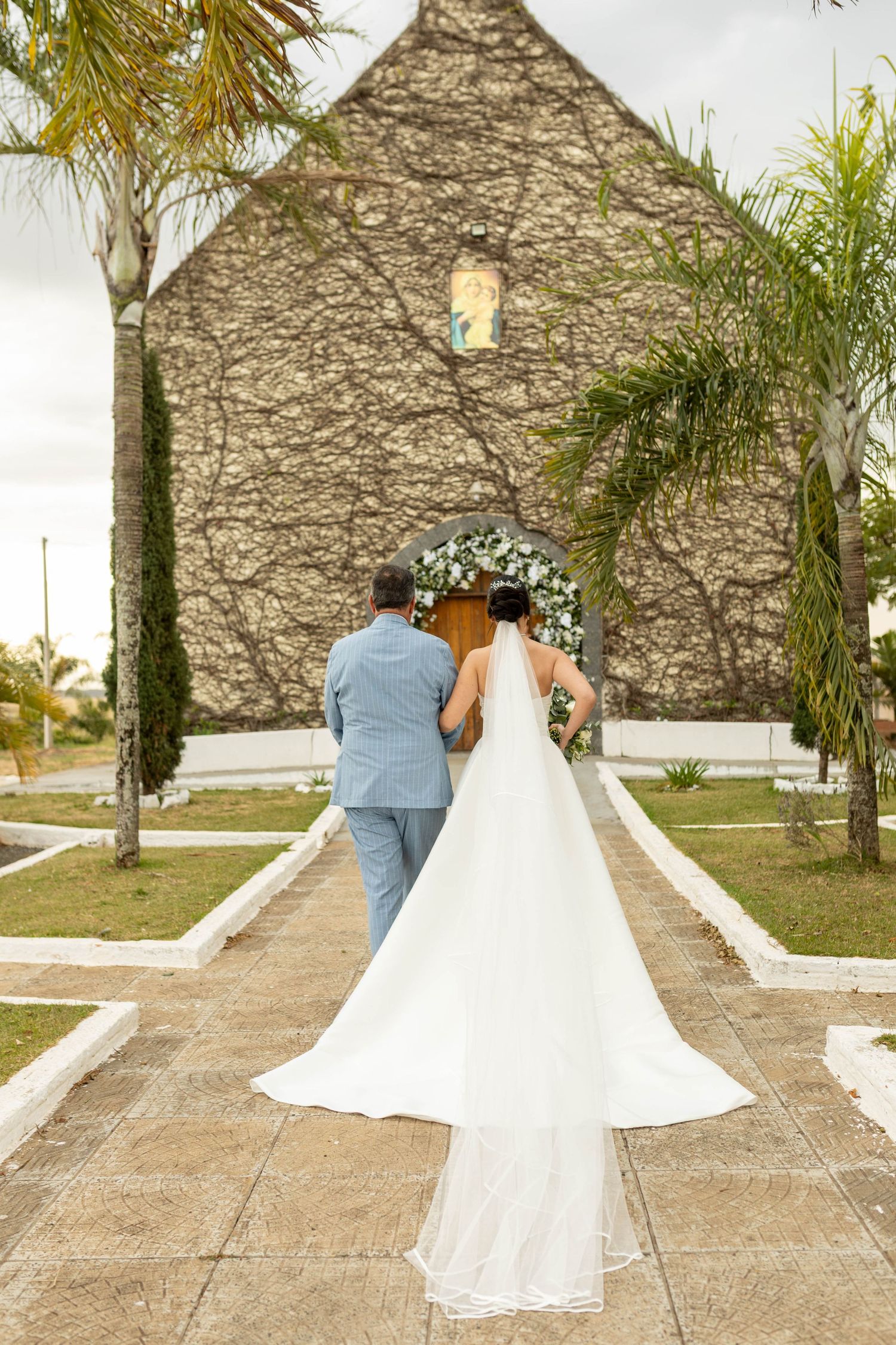 Bride in white gown with long veil walks with groom toward stone church entrance adorned with floral arch.