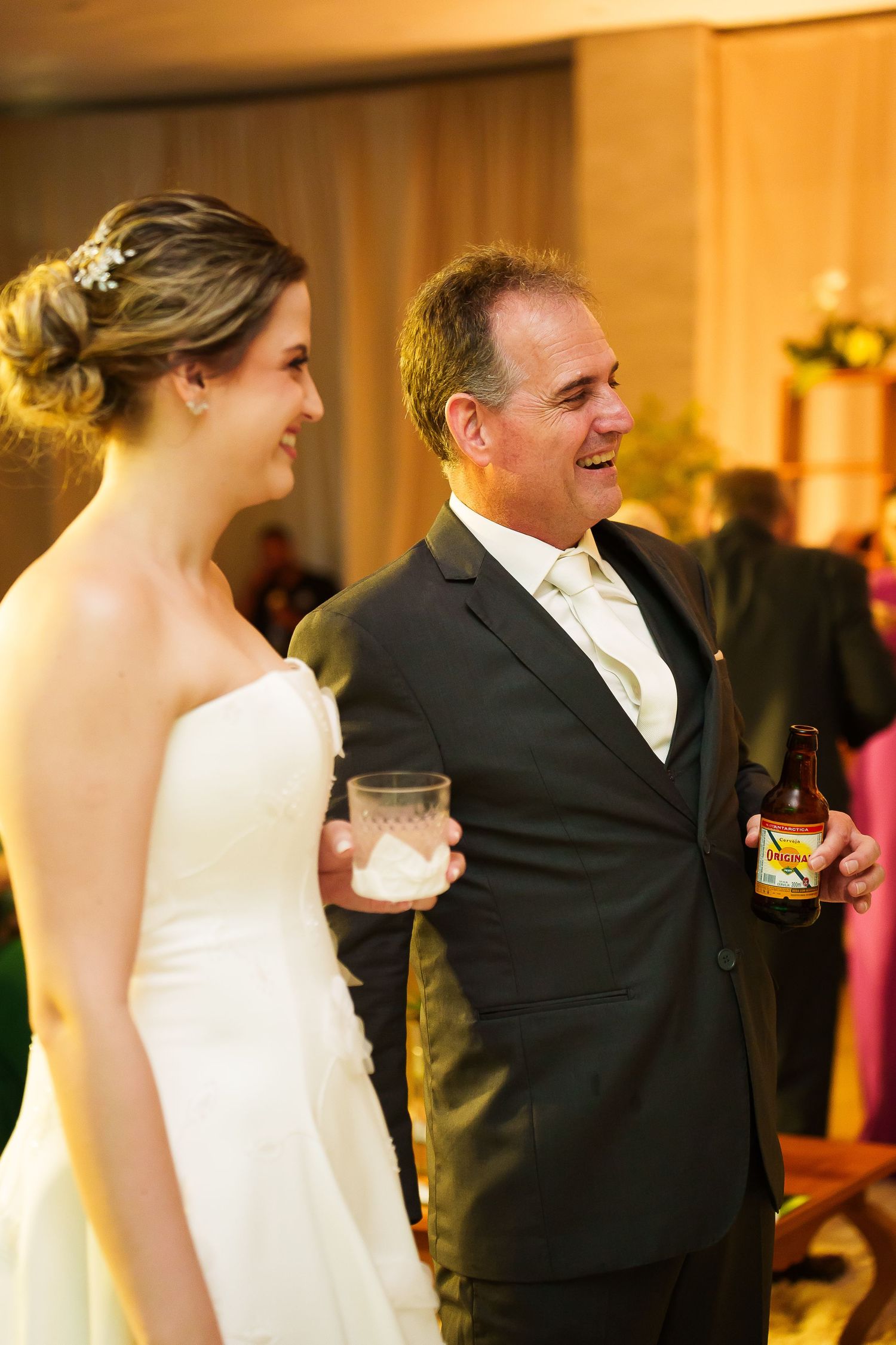 Bride in white gown and older man in dark suit holding beer bottle share a joyful moment at a wedding reception.