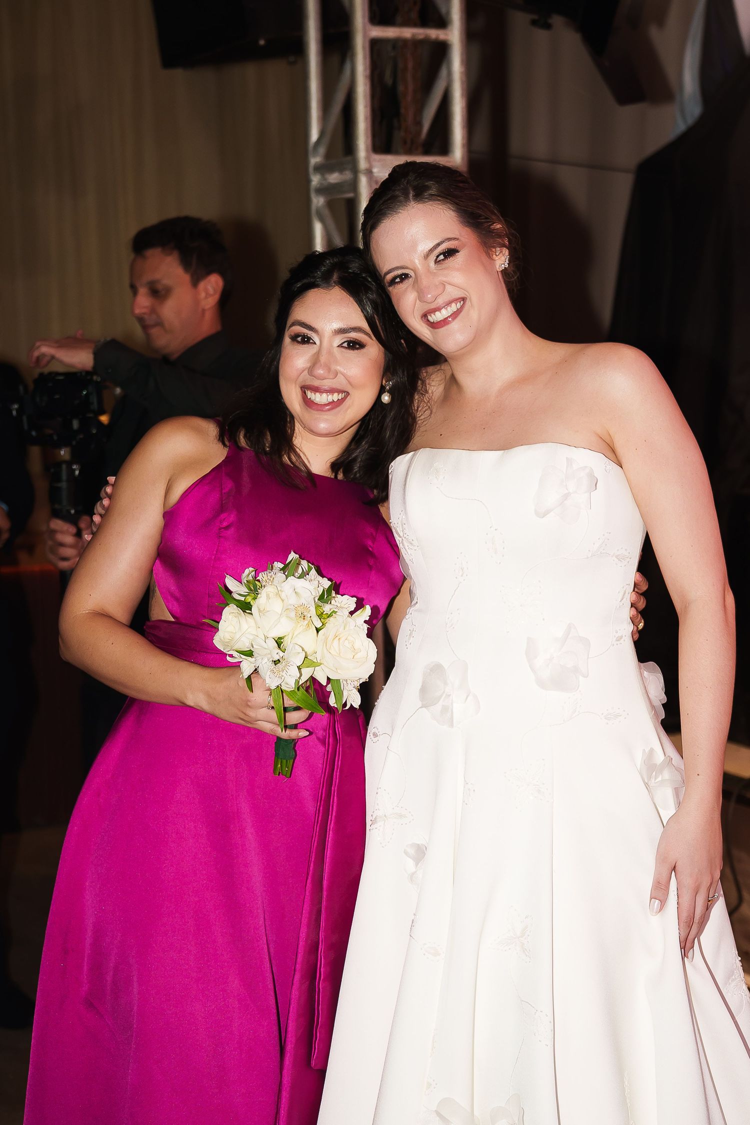 Bride in white strapless gown poses with bridesmaid in fuchsia dress holding white bouquet at wedding reception.