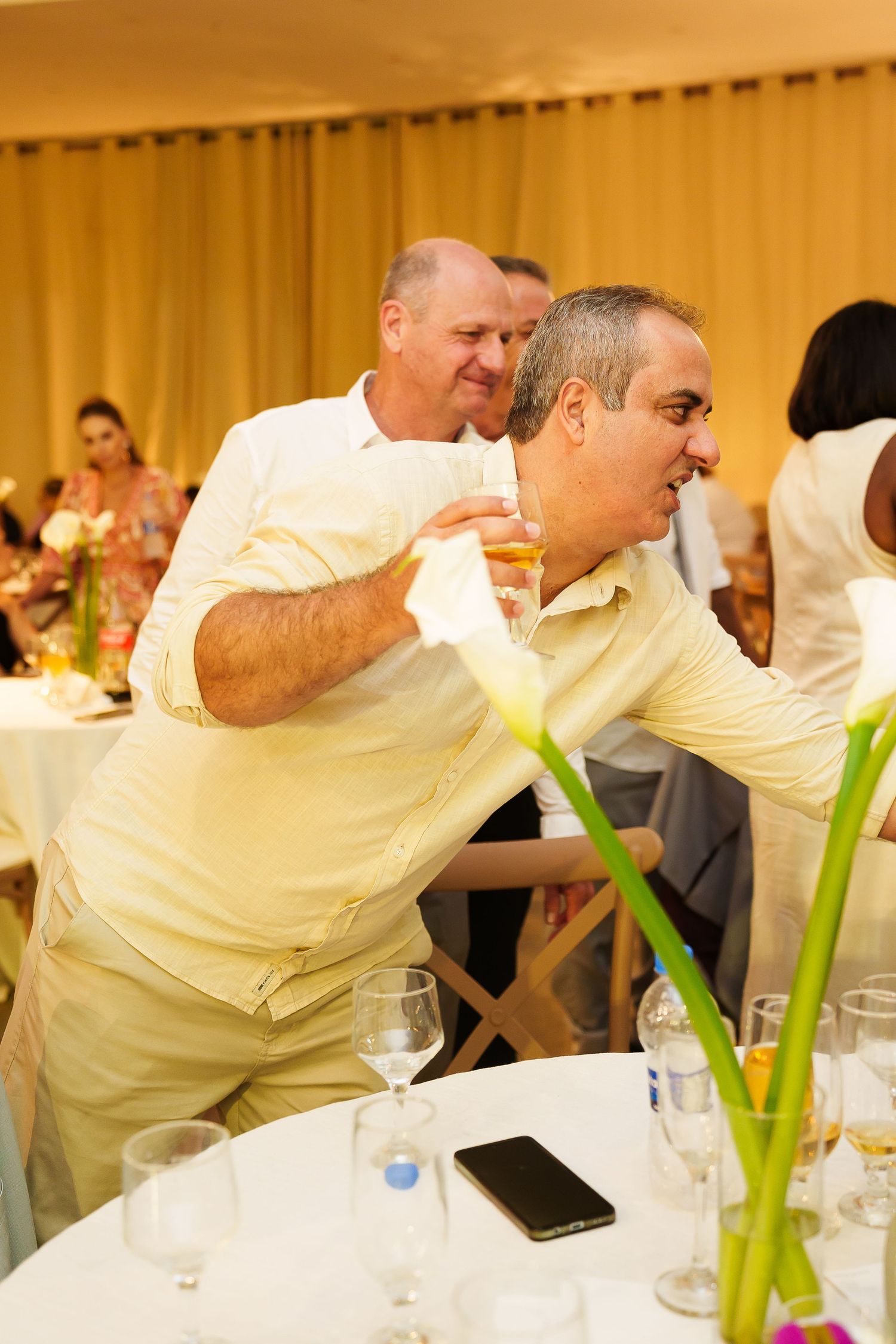 Two men in light-colored shirts socializing at an elegant indoor event, one holding a drink near a table with glassware.