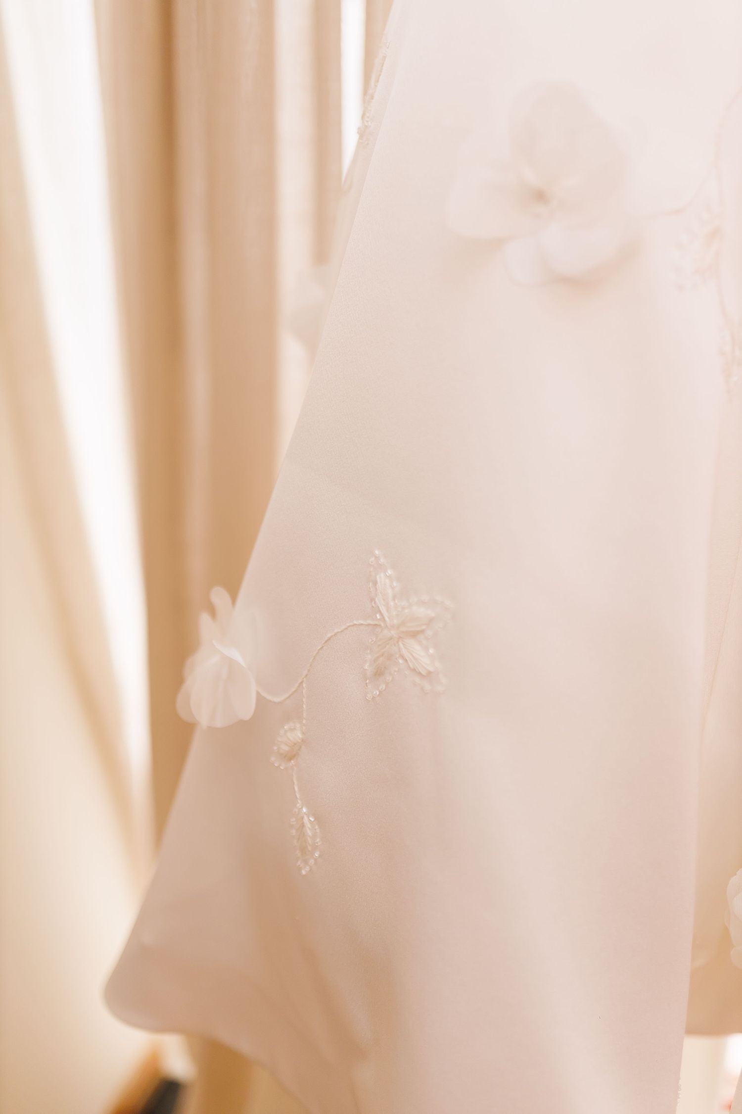 Close-up of a white wedding dress with delicate 3D floral appliques and embroidery details hanging near a window.