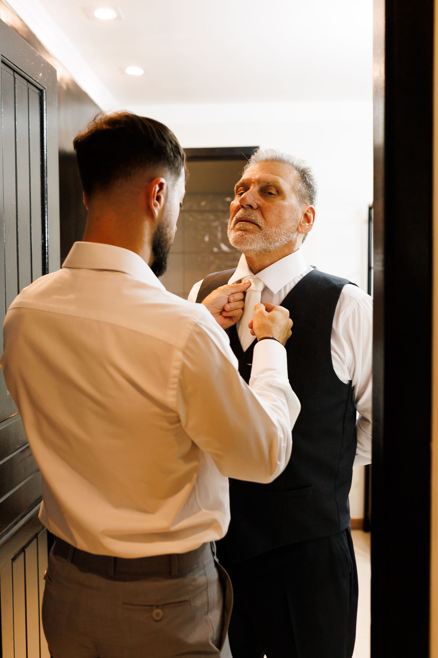 Young man helping an older gentleman adjust his tie while getting dressed in formal attire indoors.