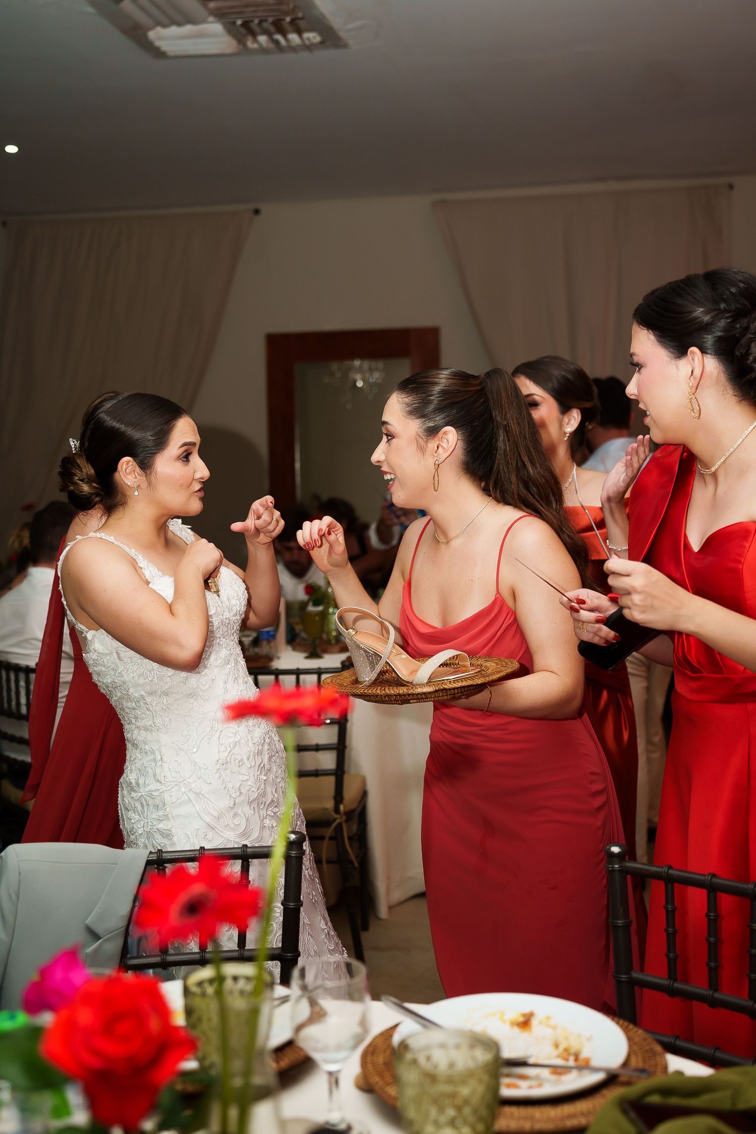 Bride in white lace gown chatting with bridesmaids in red dresses at a wedding reception dinner.
