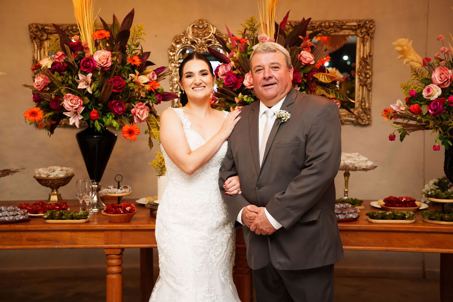 Bride in white gown poses with older man in suit before floral arrangements at elegant wedding reception.