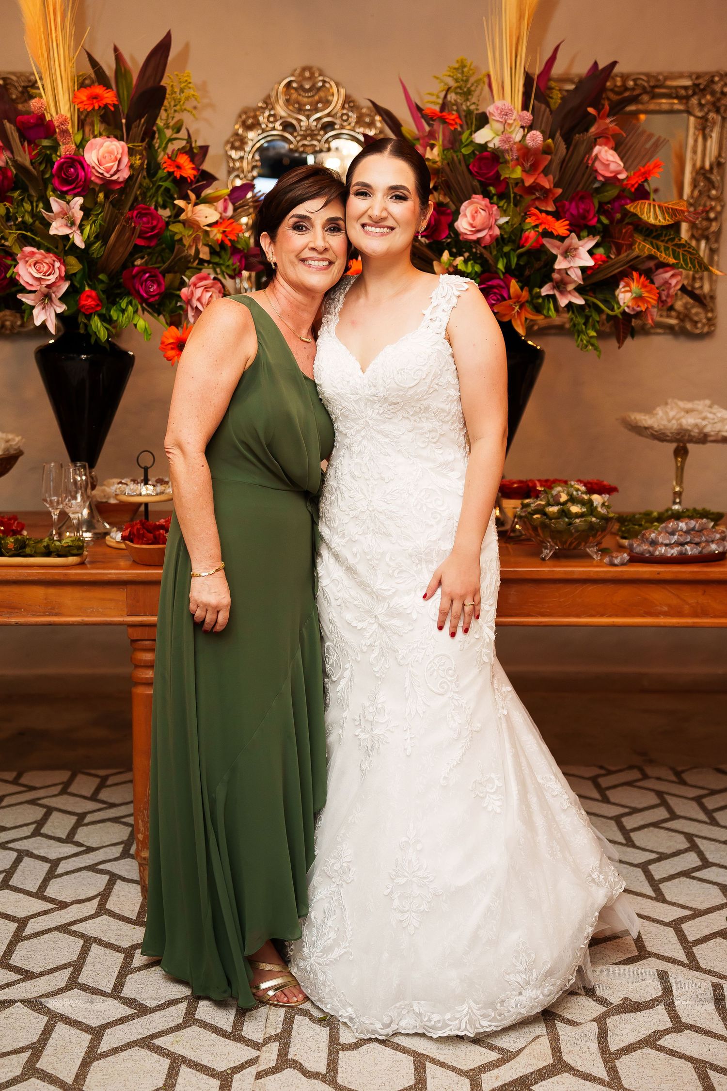 Bride in white lace gown poses with woman in green dress at elegant wedding reception with floral arrangements.