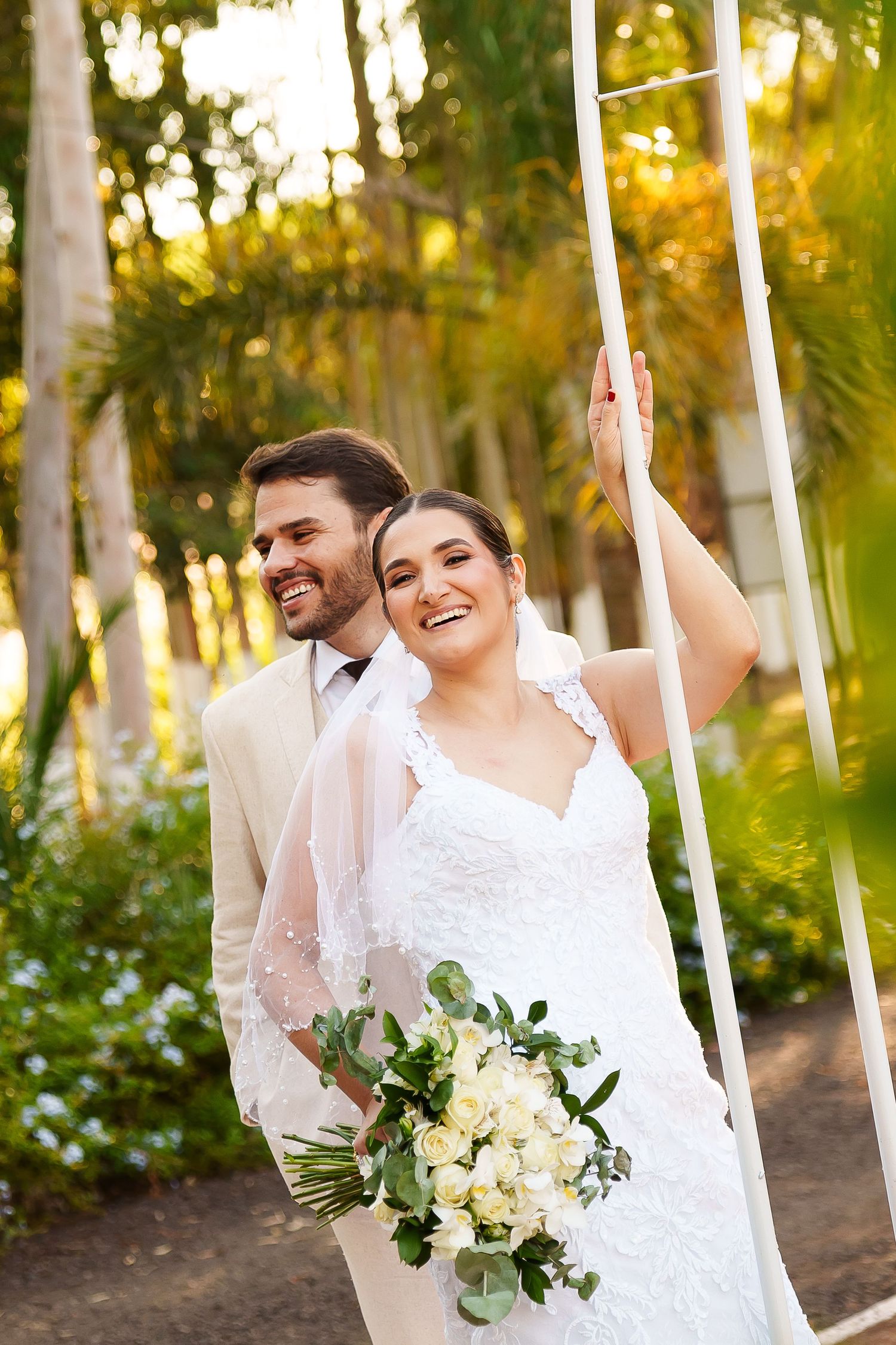 Smiling bride holding white roses bouquet with groom in cream suit, posing outdoors near palm trees on their wedding day.