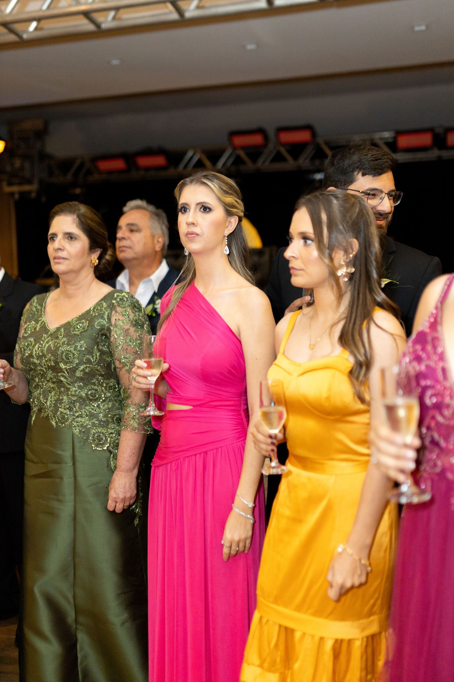 Elegant women in colorful formal gowns holding champagne glasses at an indoor event or wedding reception.