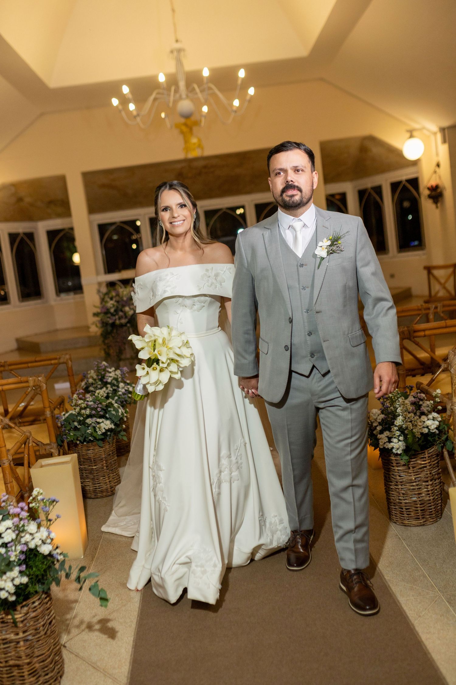Bride in off-shoulder white gown and groom in gray suit walk down church aisle adorned with floral basket arrangements.
