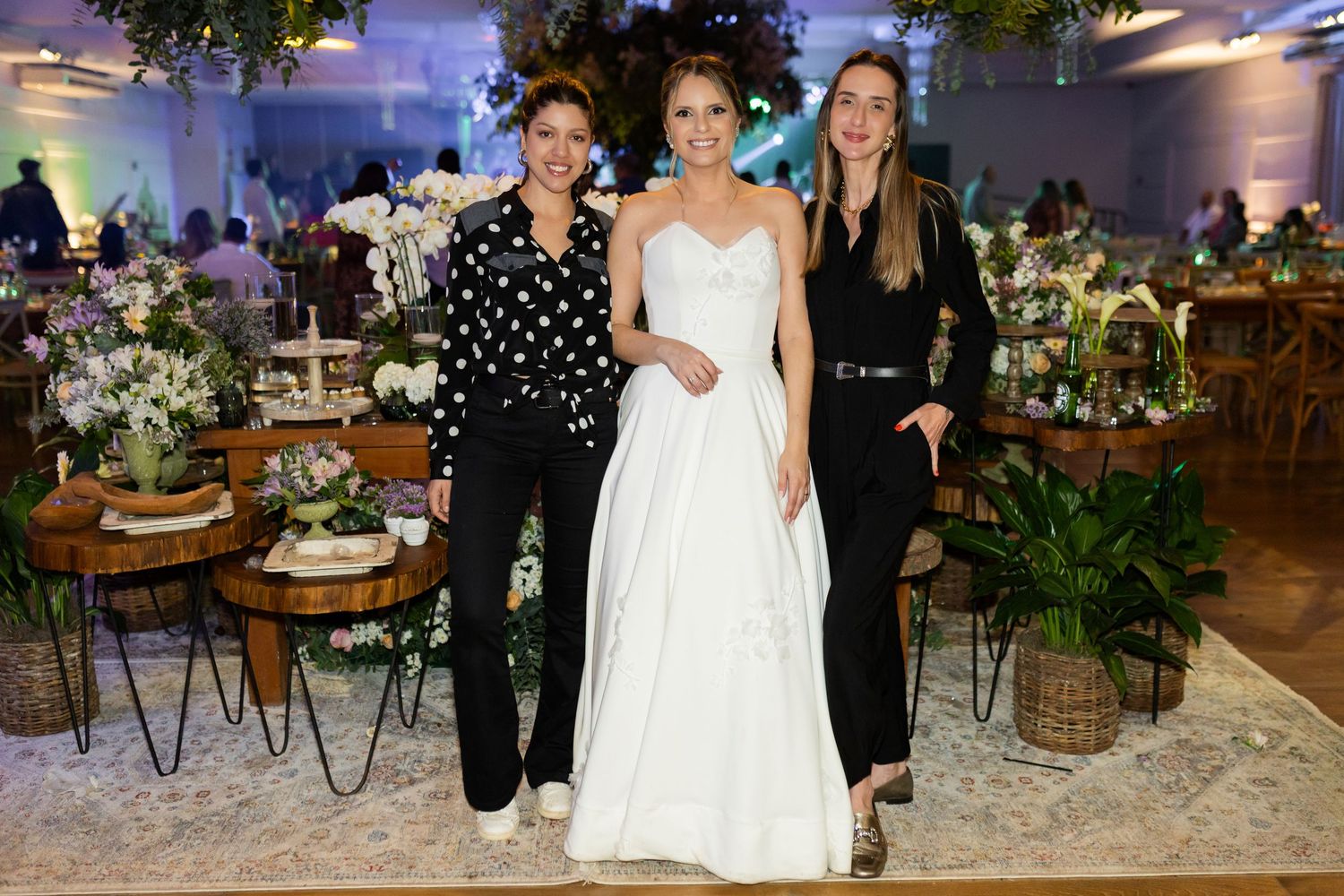Bride in white gown poses with two women in black outfits at an elegantly decorated wedding reception venue.