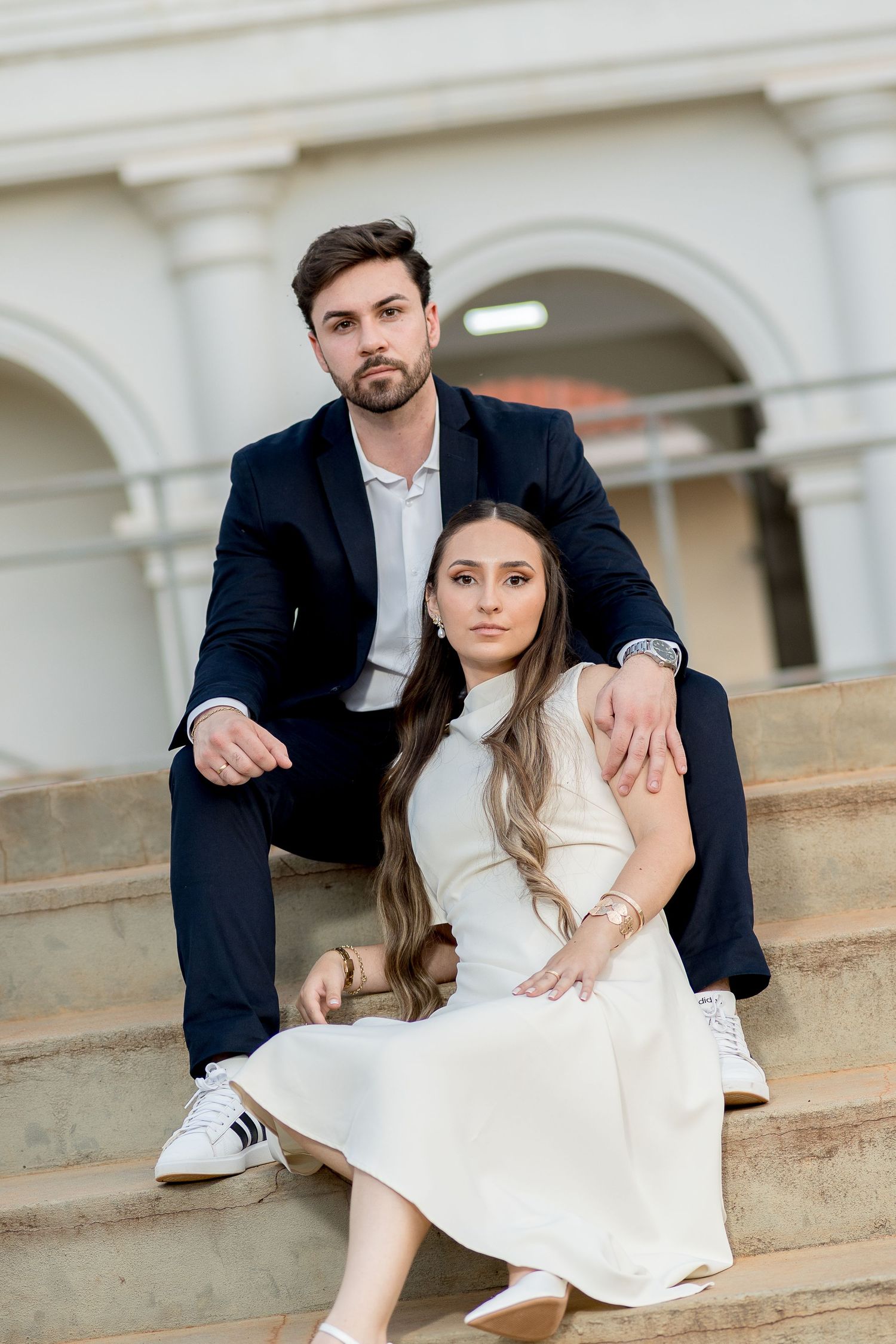 A couple in formal attire sits on stone steps, man in navy suit behind woman in white dress, posing elegantly outdoors.