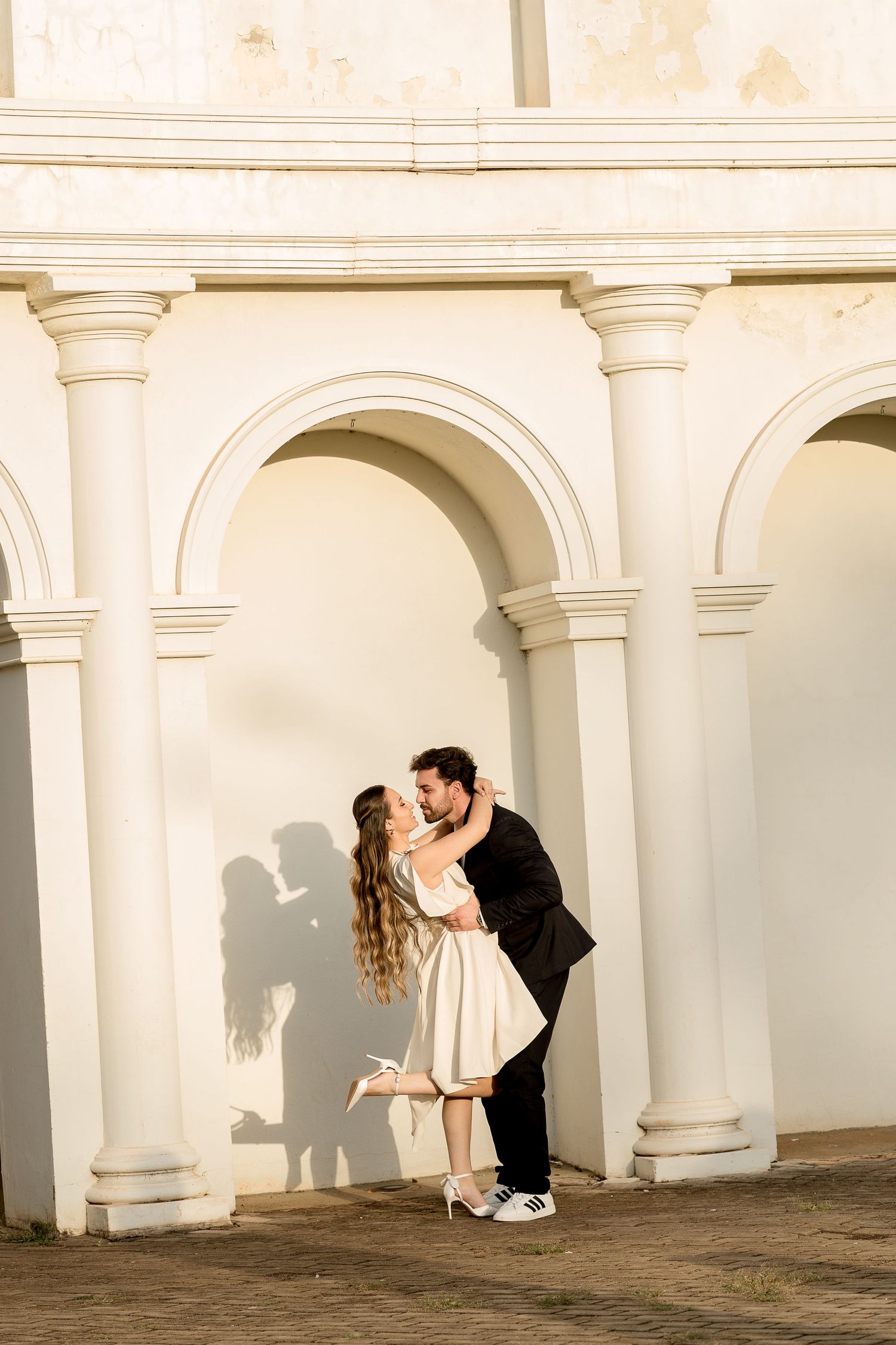 Romantic couple sharing a kiss under white arched columns, casting shadows on a sunlit classical building exterior.