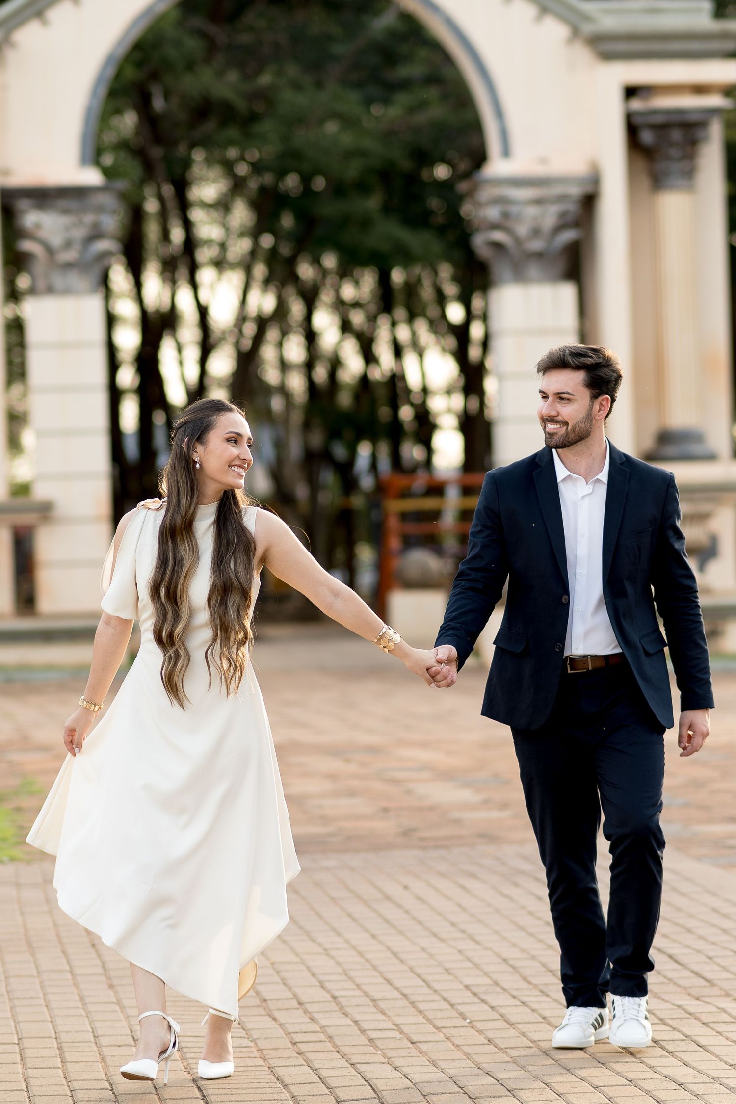 Smiling couple holding hands walking outdoors, woman in white dress and man in navy suit, elegant arch in background.