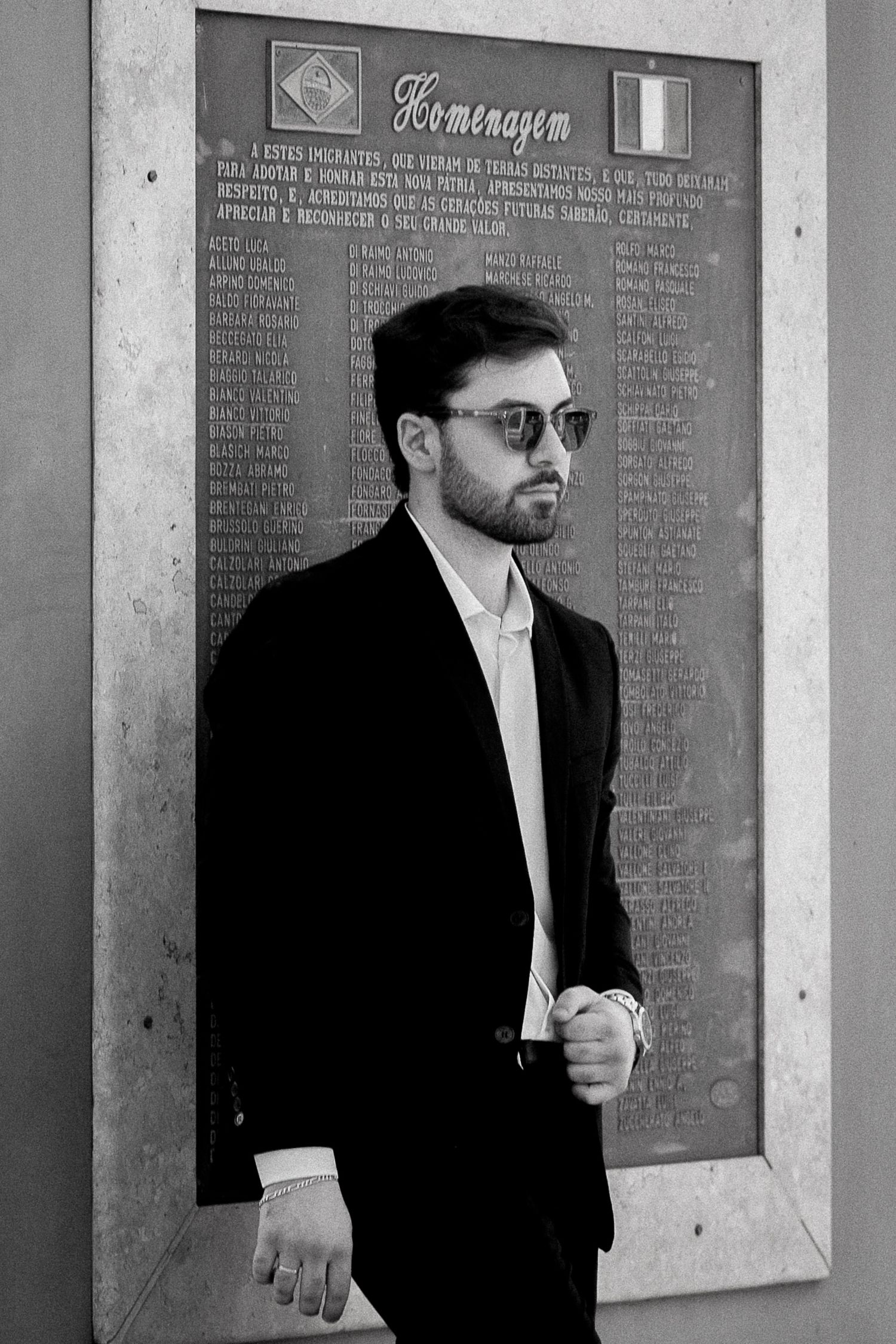 Black and white portrait of a bearded man in a suit and sunglasses standing before a memorial plaque.