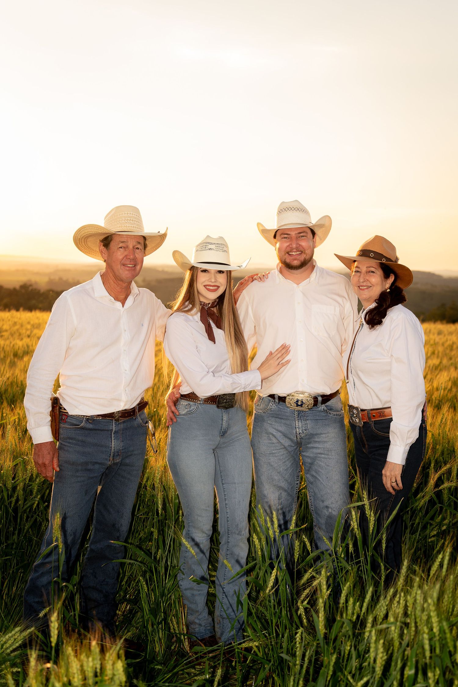 Four people in white shirts, cowboy hats, and jeans pose together in a golden wheat field at sunset.