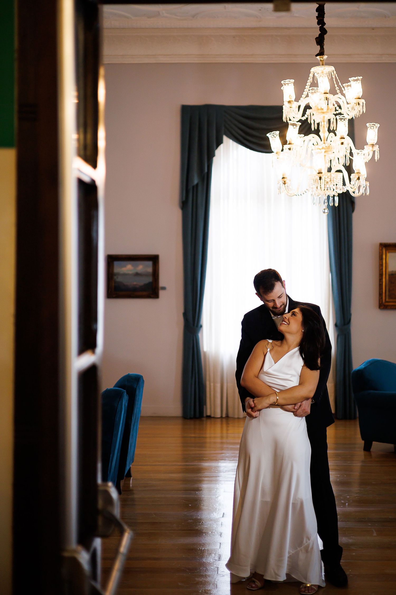 Elegant couple embracing under crystal chandelier in grand room with teal drapes, bride in white gown and groom in black suit.