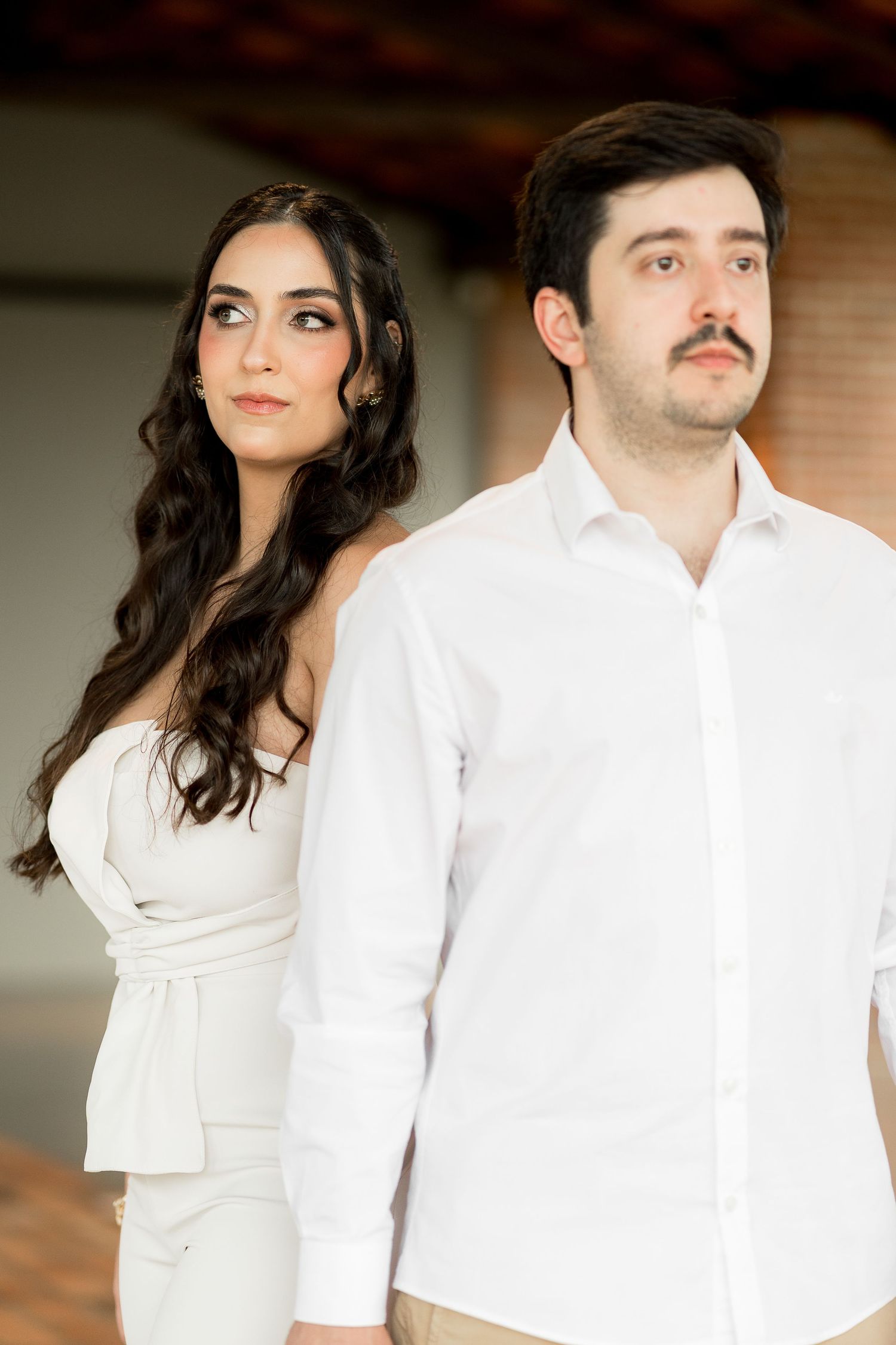 Elegant couple in white outfits posing indoors, woman with long dark hair and man with mustache in white shirt.