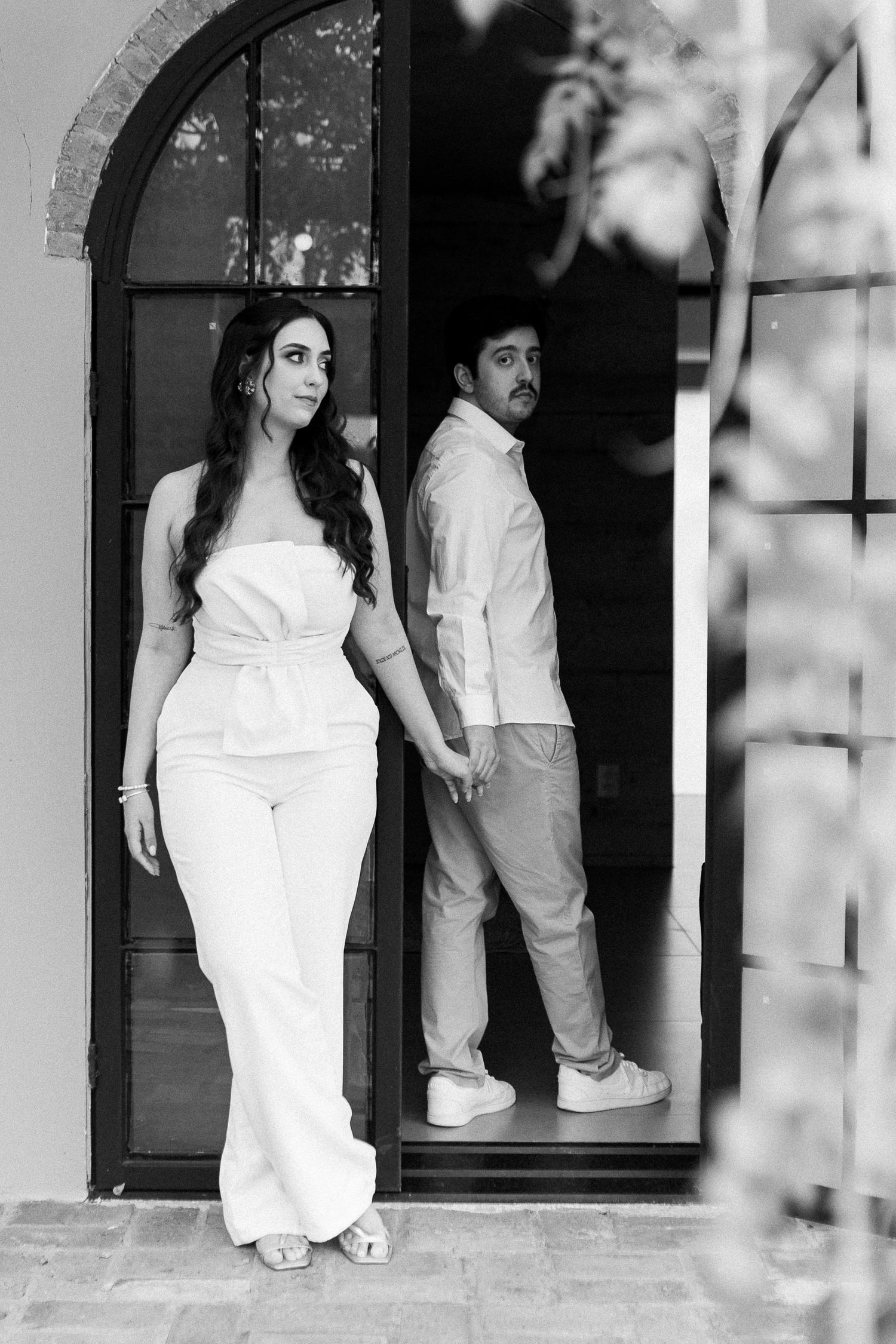 Black and white photo of a couple posing in an arched doorway, woman in white jumpsuit, man in casual attire.