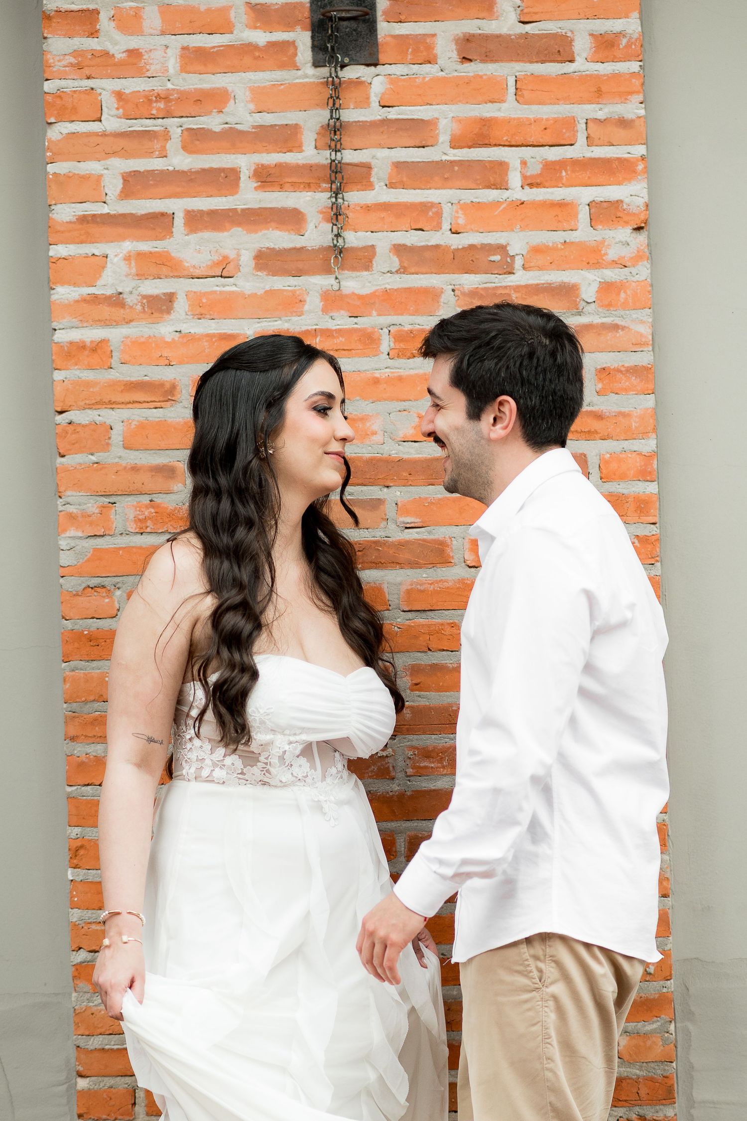 A couple in white wedding attire gazes lovingly at each other against a rustic red brick wall.