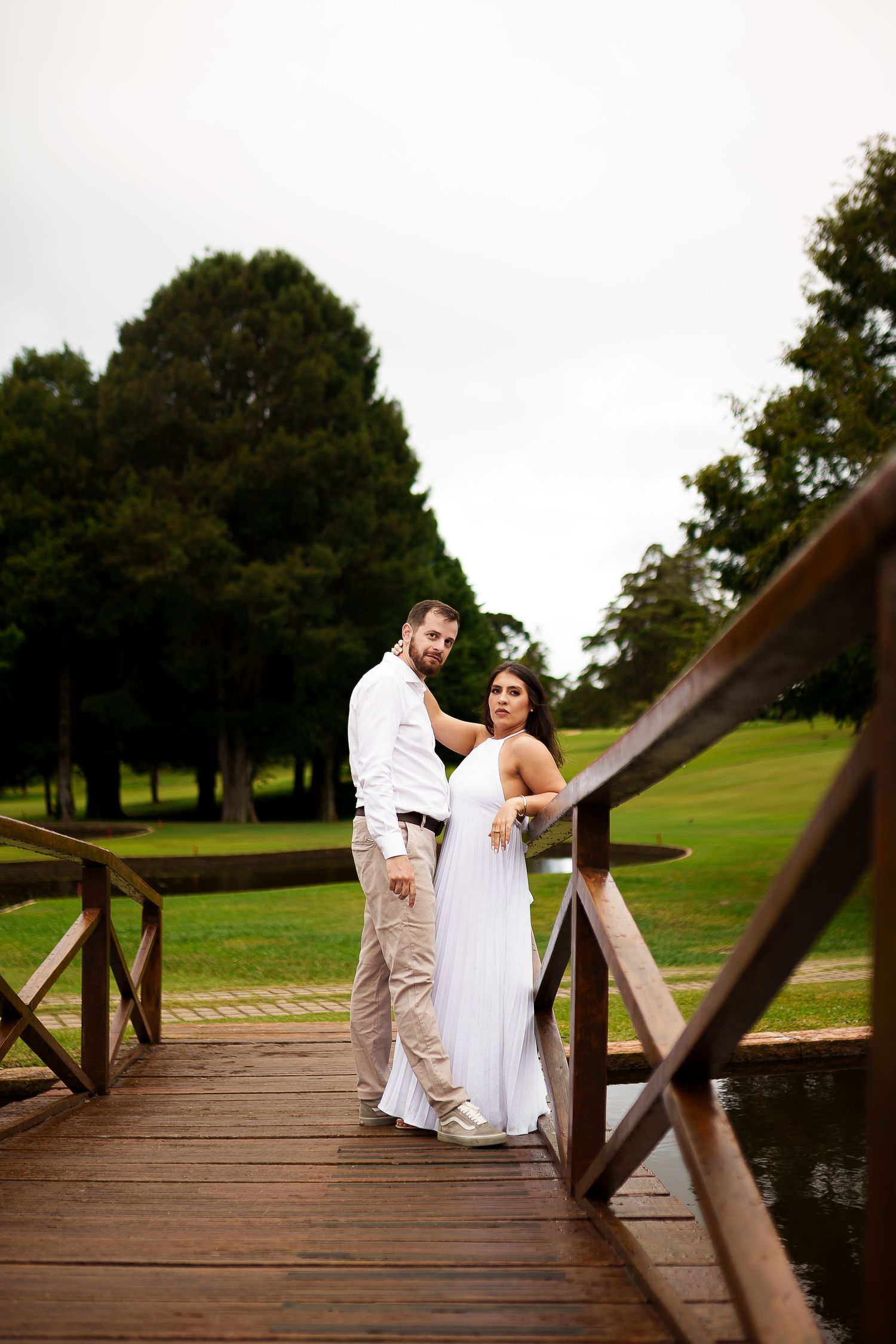 Couple in wedding attire posing on a wooden bridge surrounded by lush green trees and a serene pond.