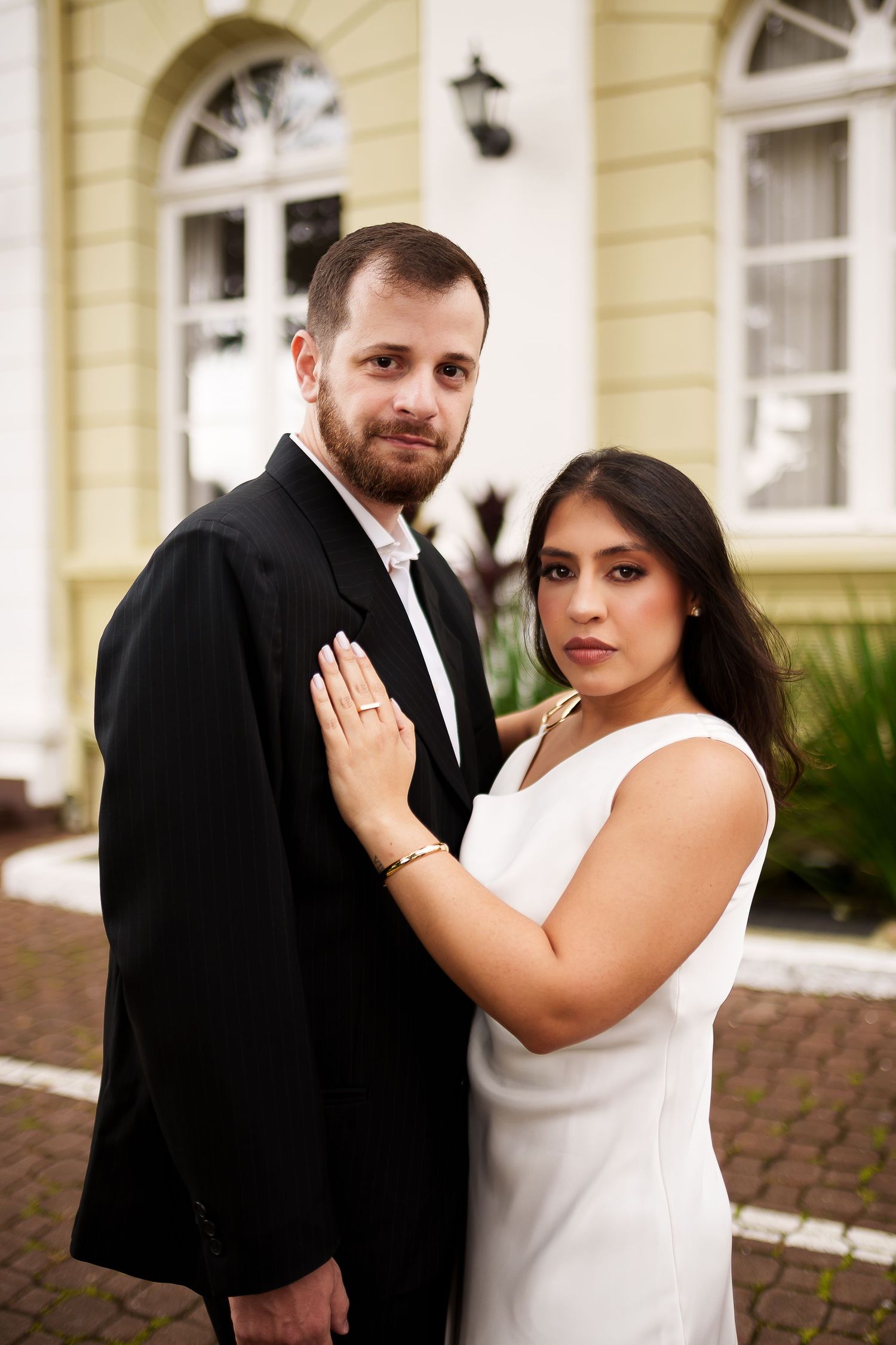 Elegant couple posing outdoors, man in black suit and woman in white dress, showcasing engagement ring near a classic building.