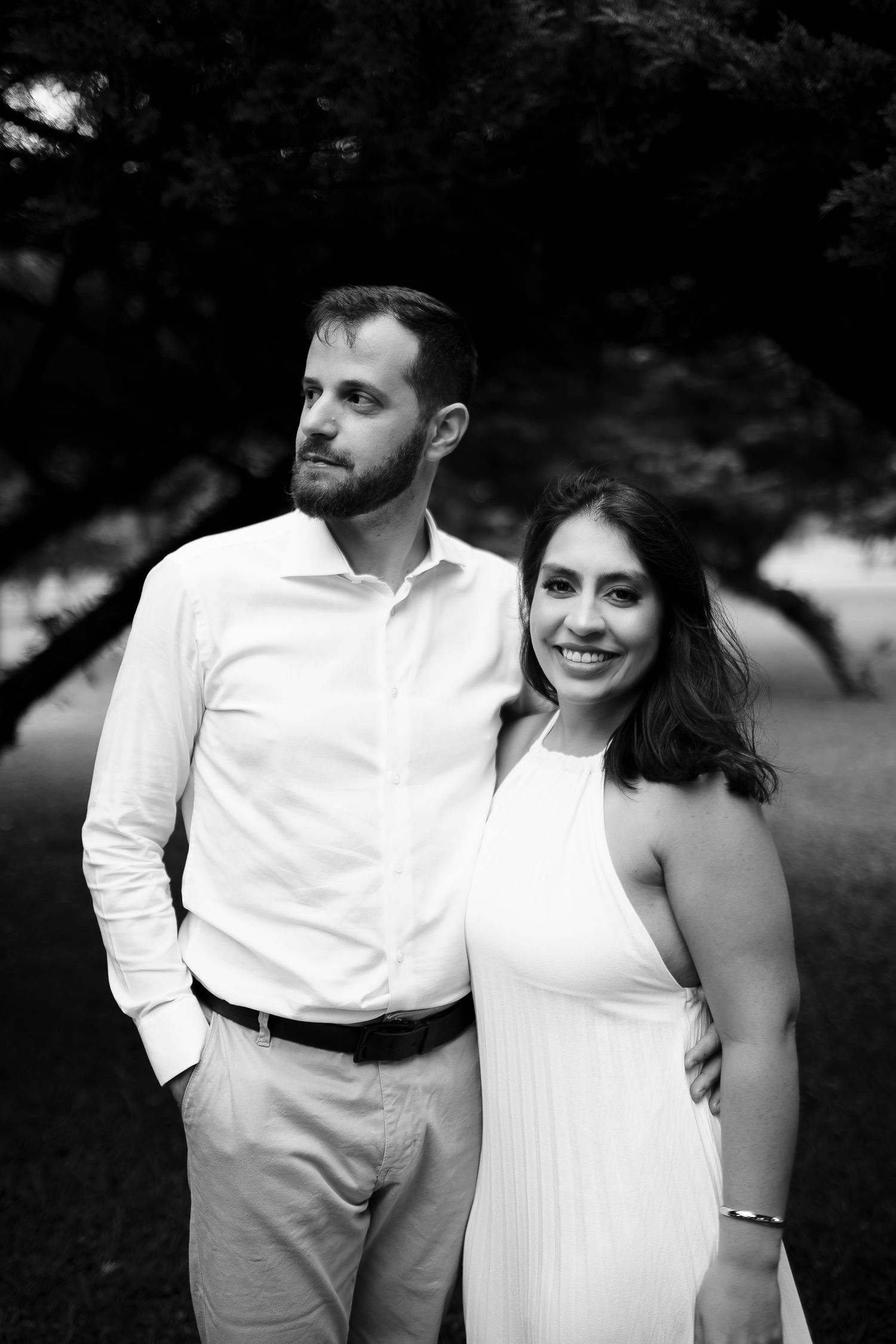 Black and white photo of a smiling couple in white outfits posing outdoors near trees.