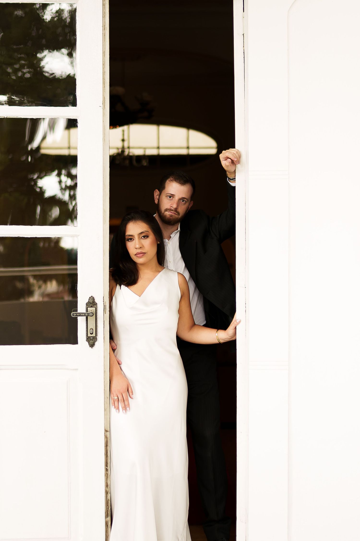 Bride in white gown and groom in black suit pose elegantly in white French doors during wedding portrait.