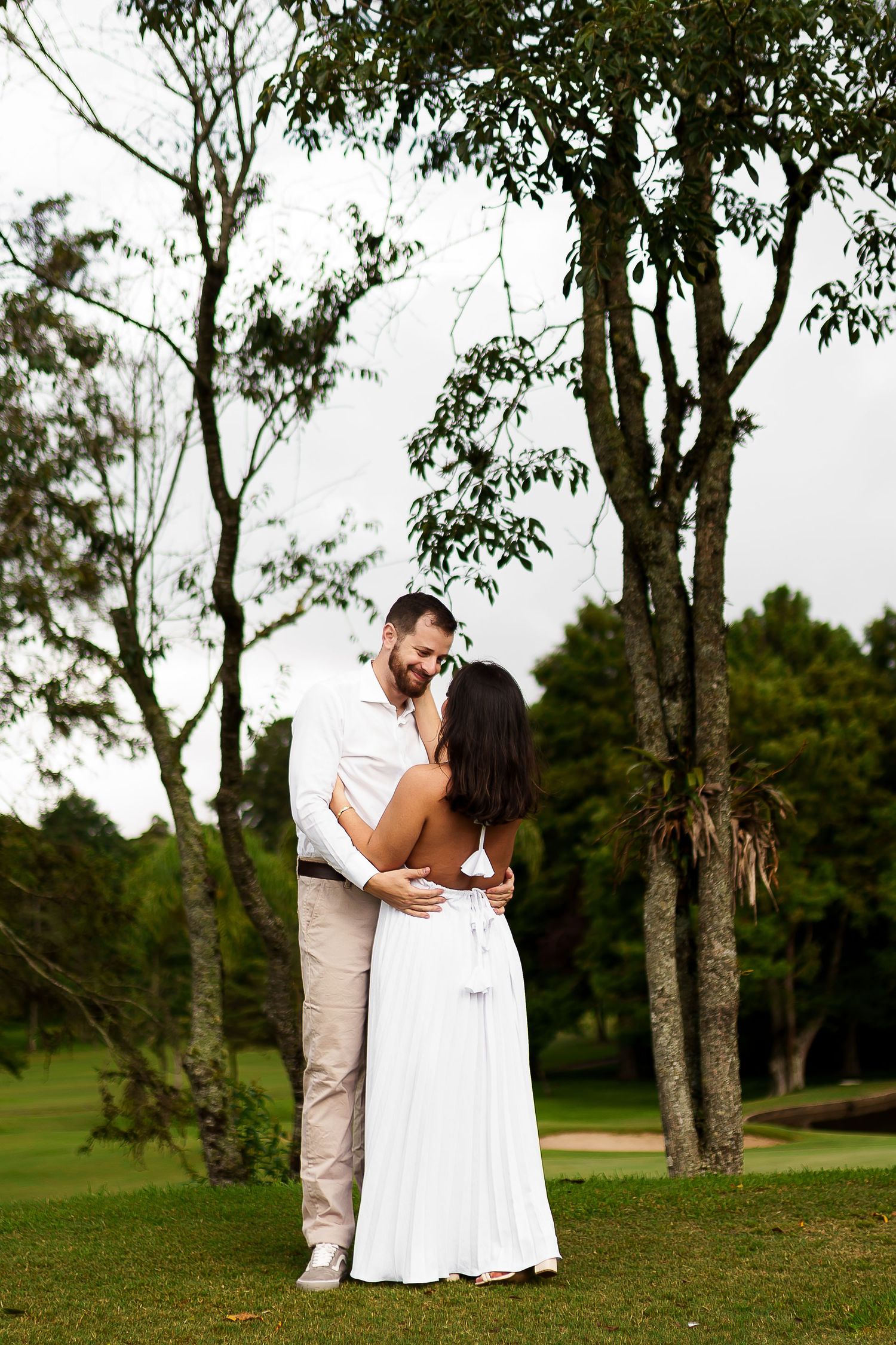 Couple in white attire embracing romantically outdoors on green grass with tall trees in background.