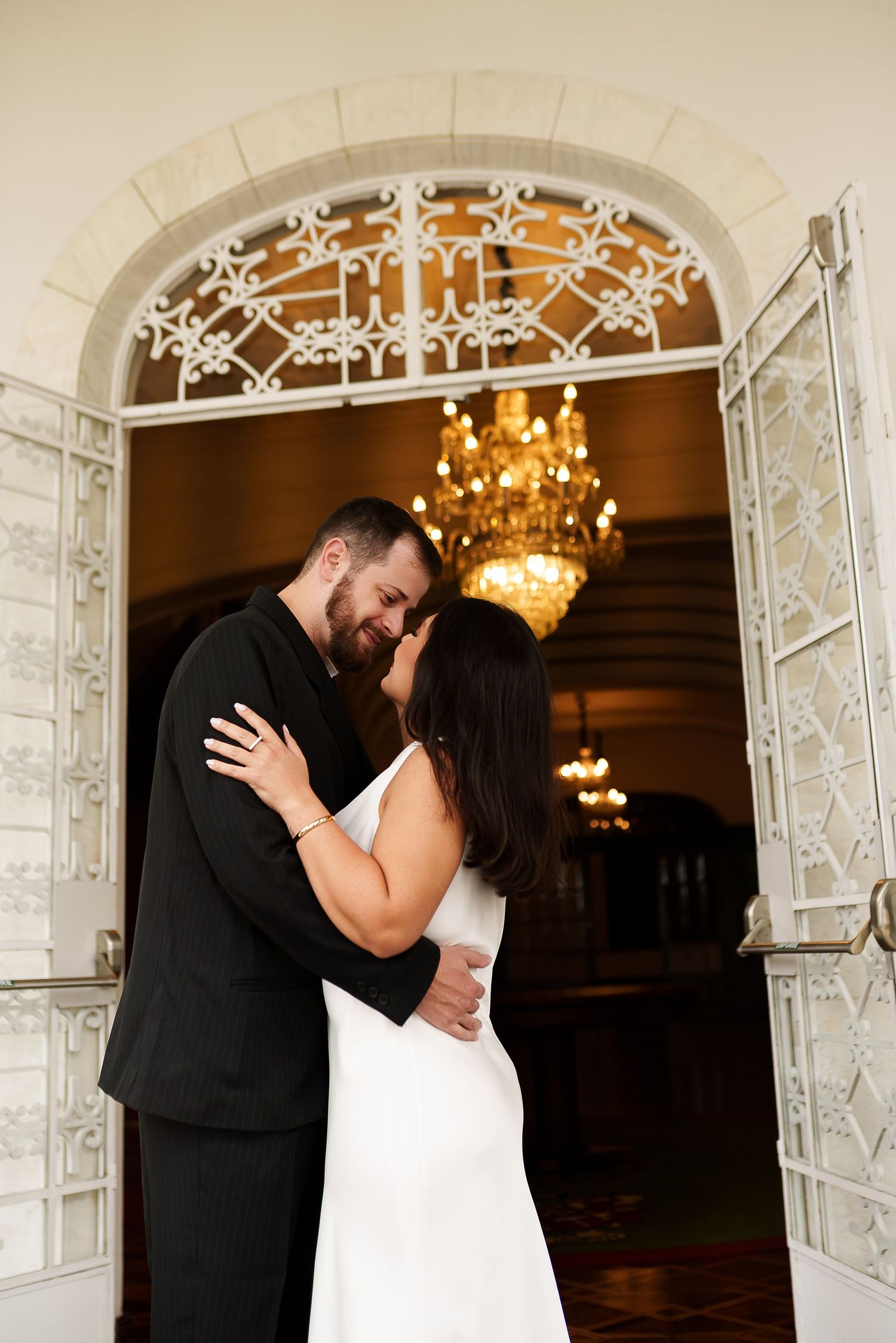 Couple sharing a romantic kiss in white wedding attire before an ornate arched doorway with glowing chandelier.