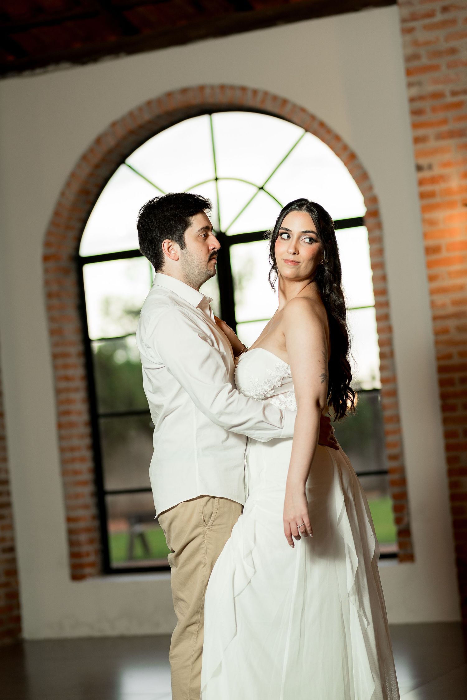A couple poses romantically in front of an arched window with brick walls, the woman in a white gown and man in a cream suit.