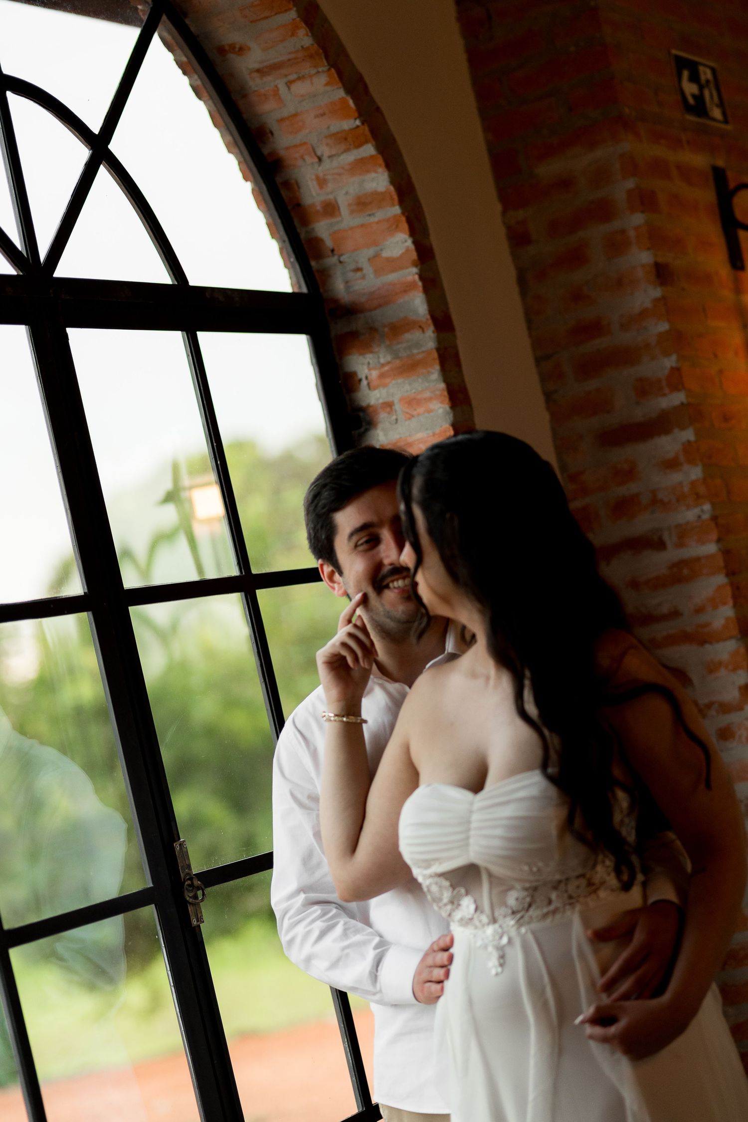 A bride and groom share a romantic moment by an arched window in a brick venue, smiling and embracing on their wedding day.