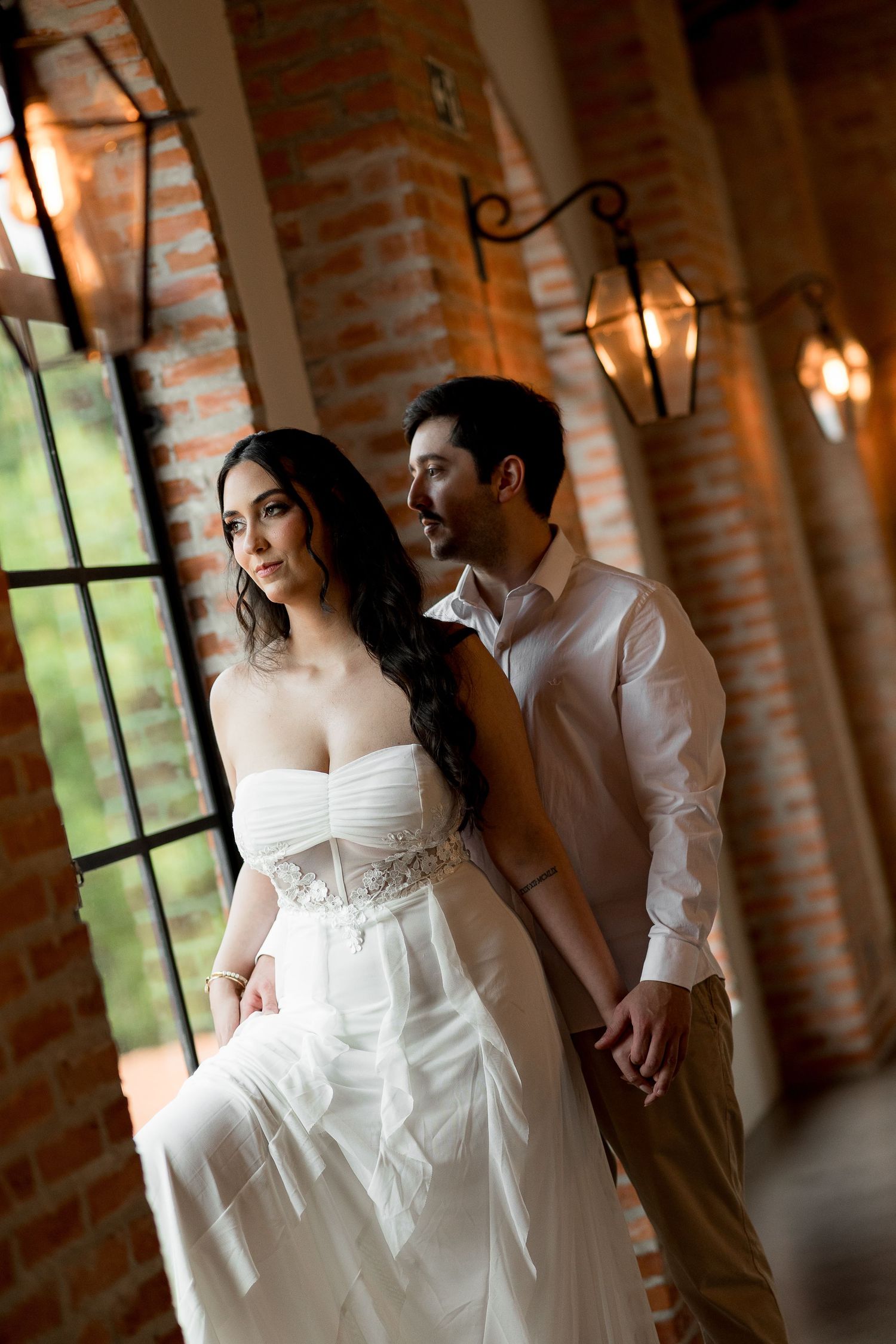 Elegant couple in wedding attire posing in a rustic brick hallway with vintage lanterns and large windows.
