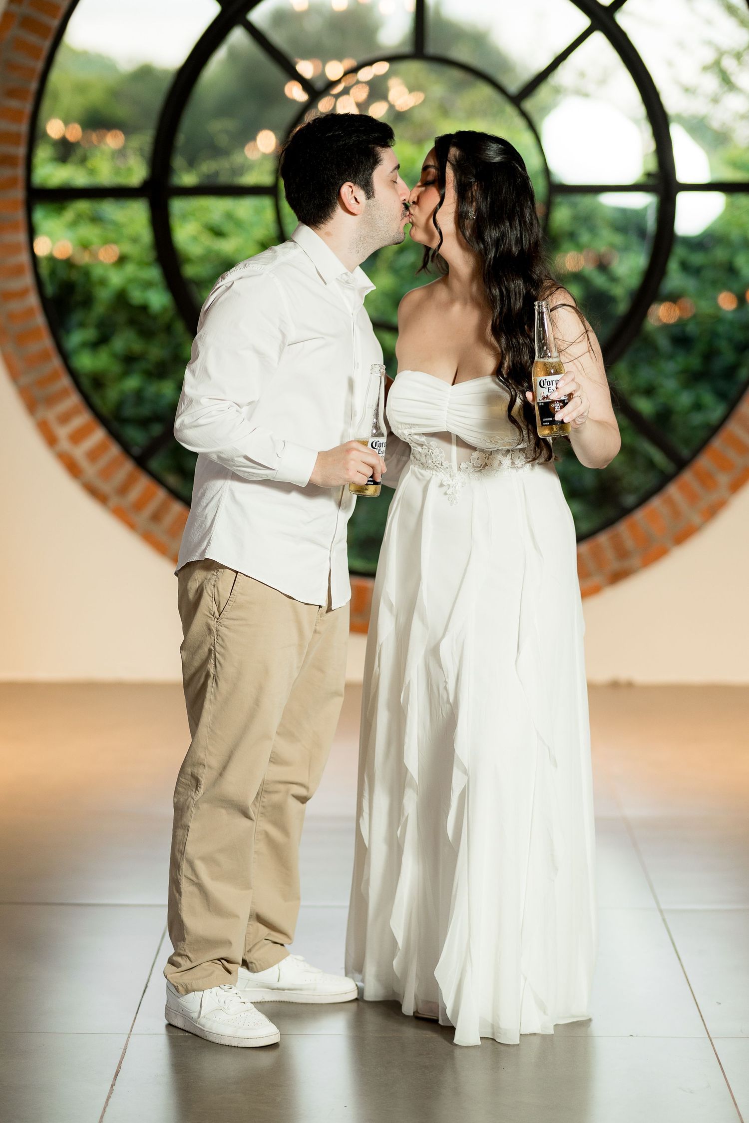 Couple sharing a romantic kiss in elegant attire, holding drinks in front of a circular window with bokeh lights.
