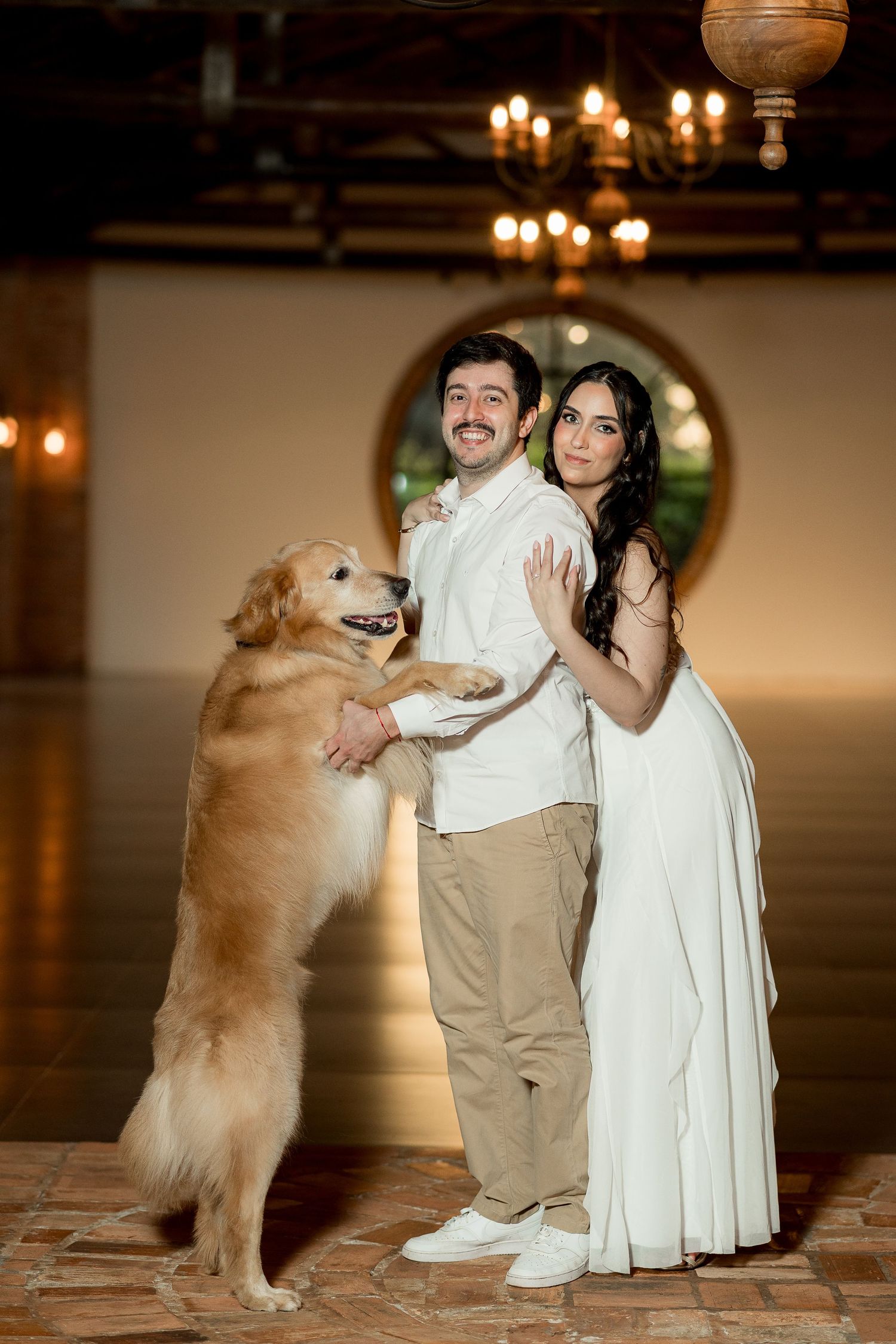 A couple in white wedding attire poses with a golden retriever standing on its hind legs inside an elegant venue.