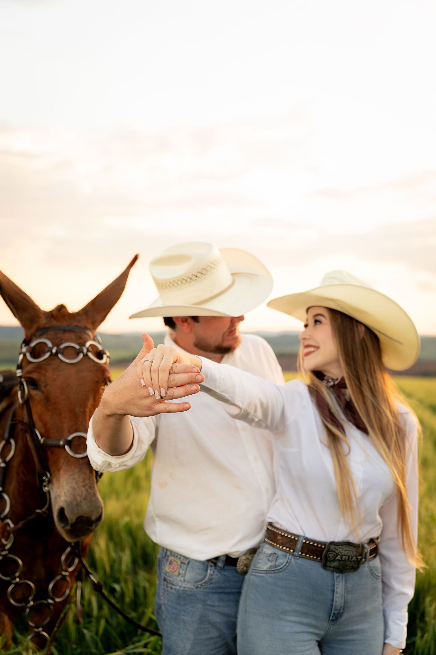 Couple in white cowboy hats and jeans showing engagement ring beside a brown horse at golden hour.